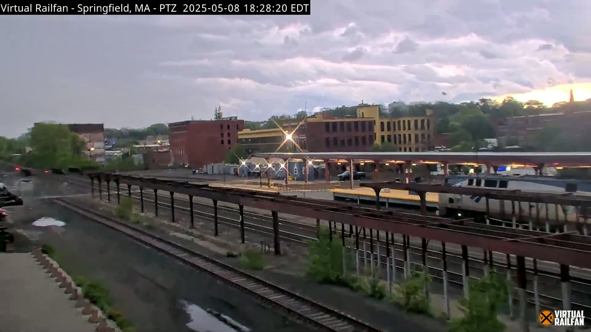 A passenger train passes through a railway station under an overcast sky.