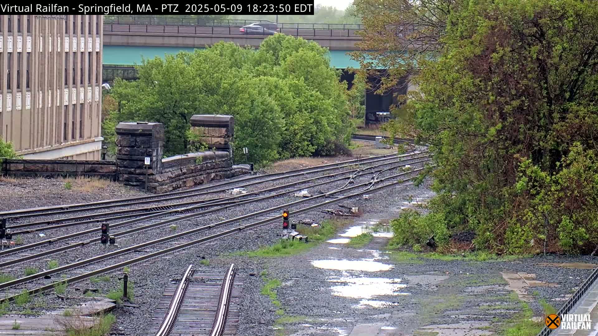 A wet, overcast day shows several railway tracks splitting and curving through a landscape of low-lying vegetation and stone structures, next to a multi-story building.