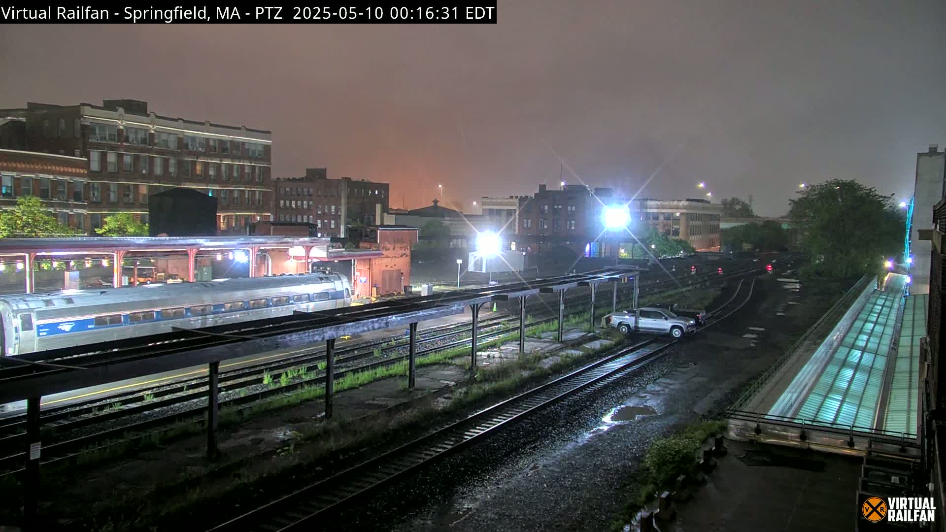 A nighttime view of a train station with a passenger train on the tracks under rainy conditions.