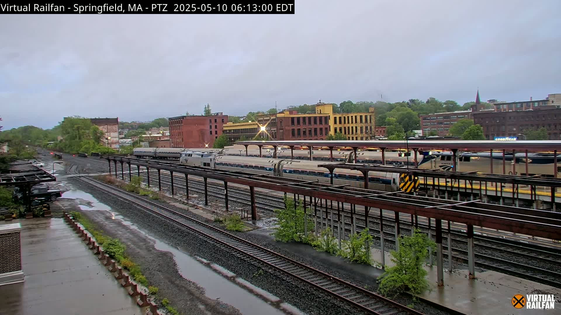 Several passenger trains are at a station platform under an overcast sky, with puddles visible on the ground.