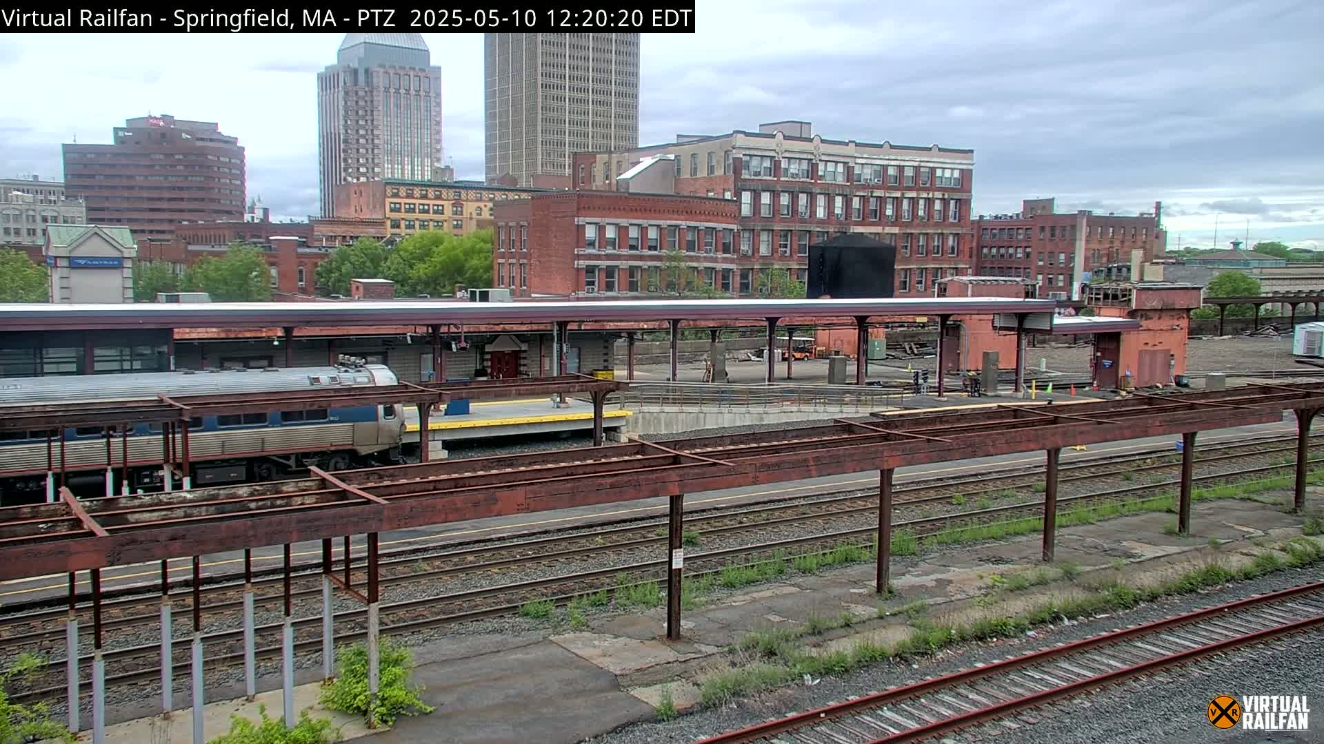 A train sits at a station platform in an urban area under an overcast sky.