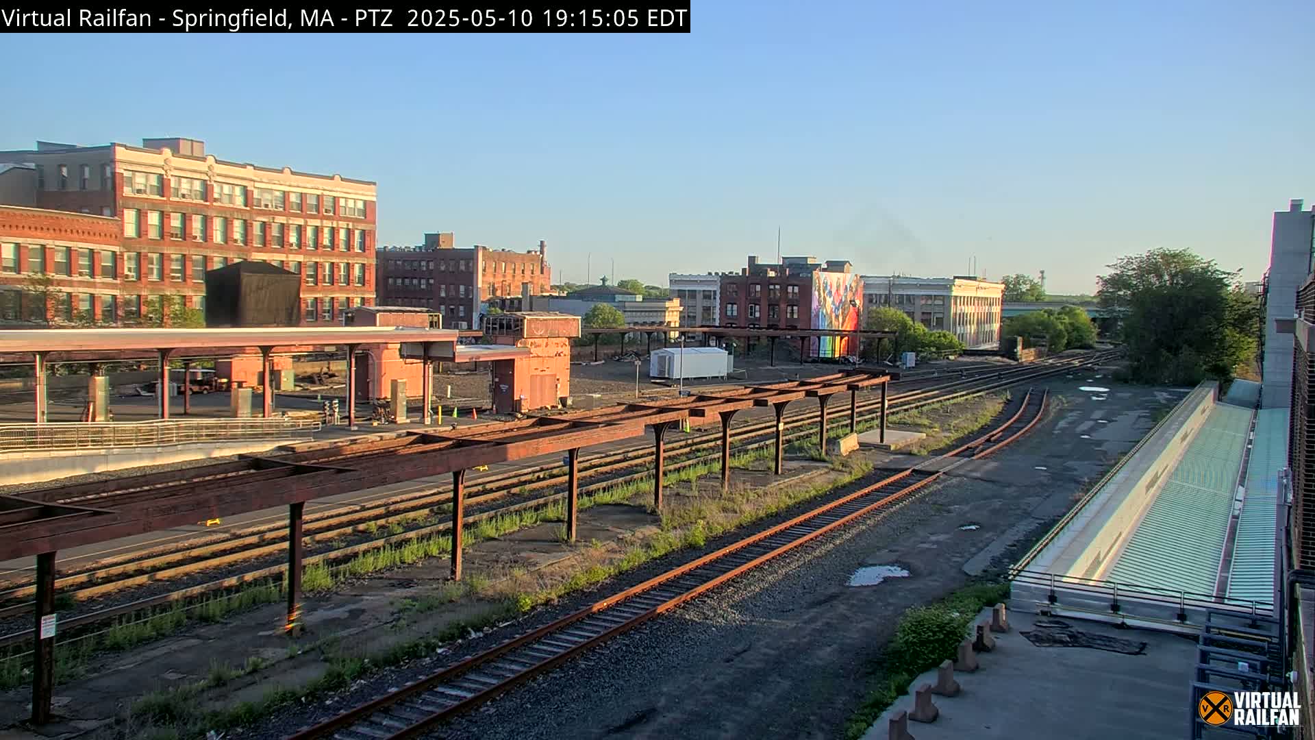 A train station platform with several railway tracks under a clear, bright sky, backed by brick buildings and a large mural.