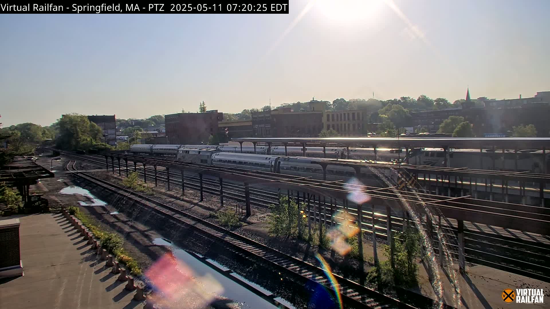 Several passenger trains are parked at a station platform under a sunny sky, with some puddles on the ground near the tracks.