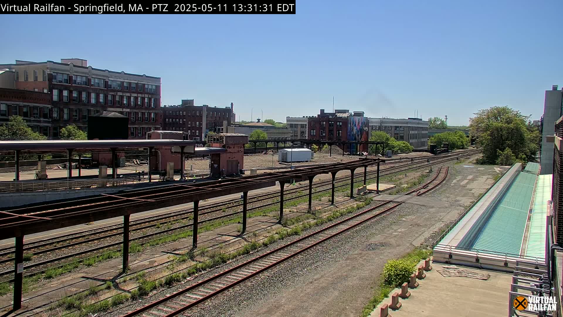 A train station with several tracks and covered platforms is visible under a clear sky, surrounded by brick buildings and overgrown vegetation.