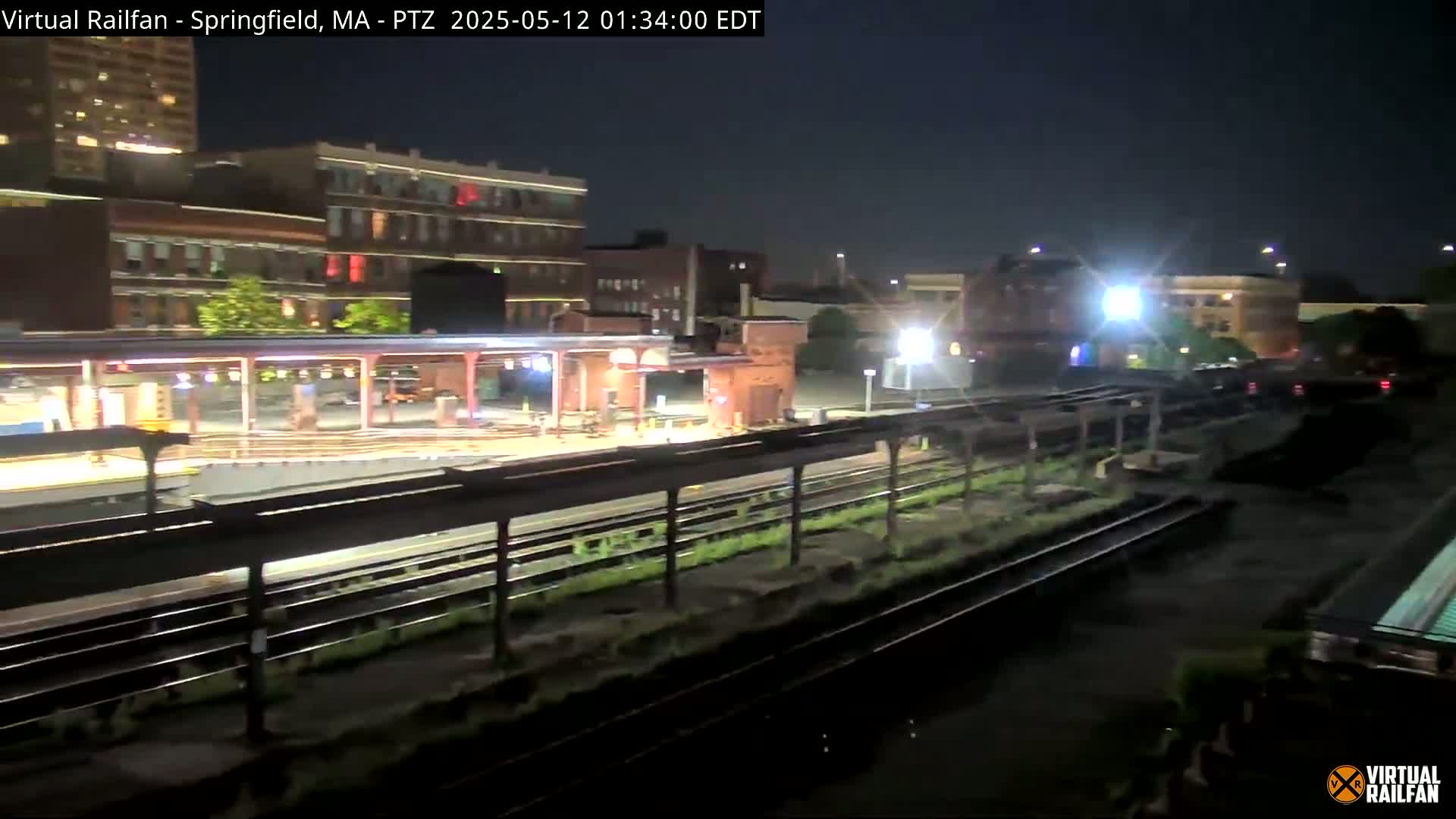 A nighttime view of a train station platform and tracks, with city buildings illuminated in the background under a clear, dark sky.