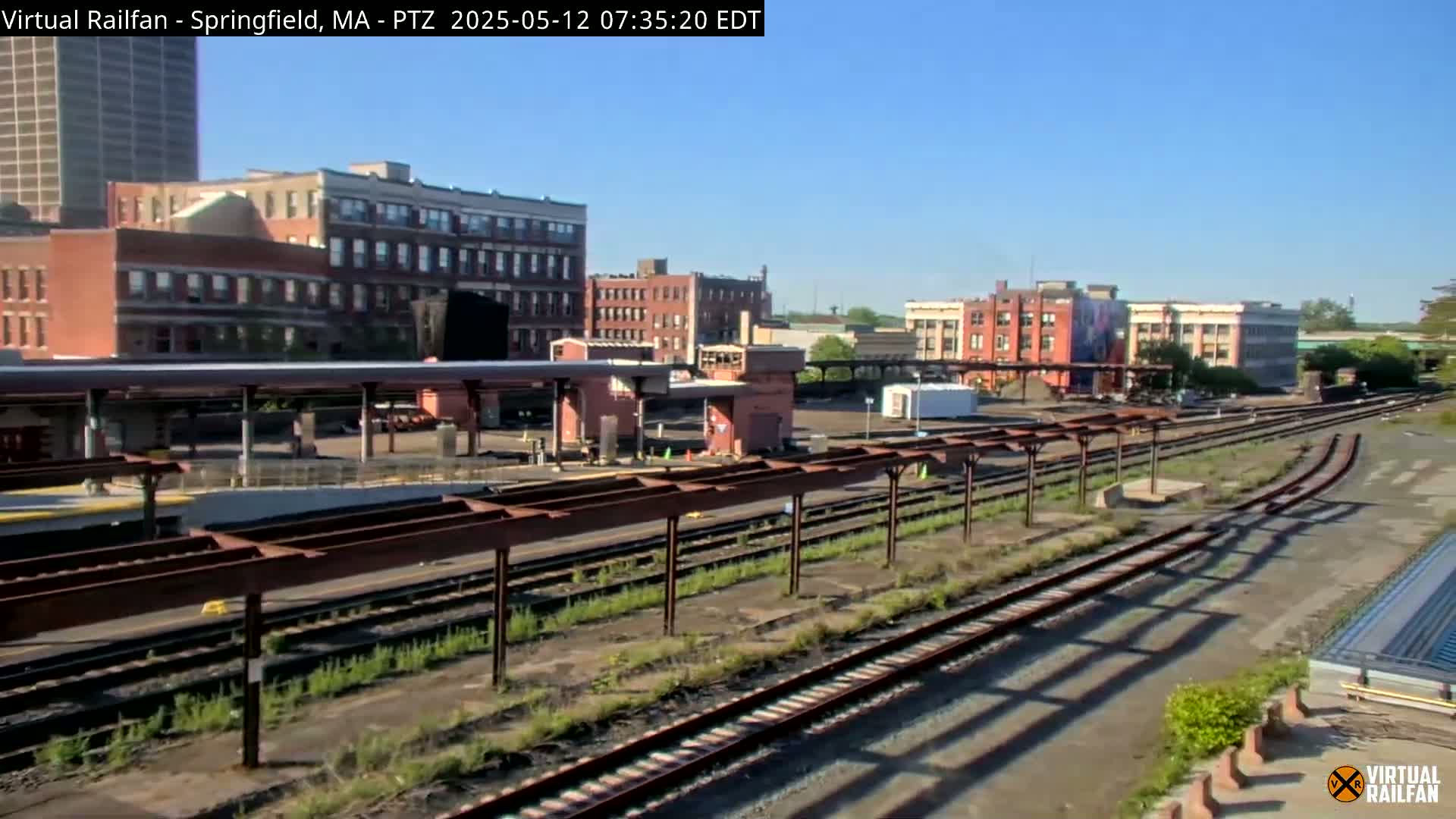 A train station with multiple tracks and platforms is situated amongst brick buildings under a clear, sunny sky.