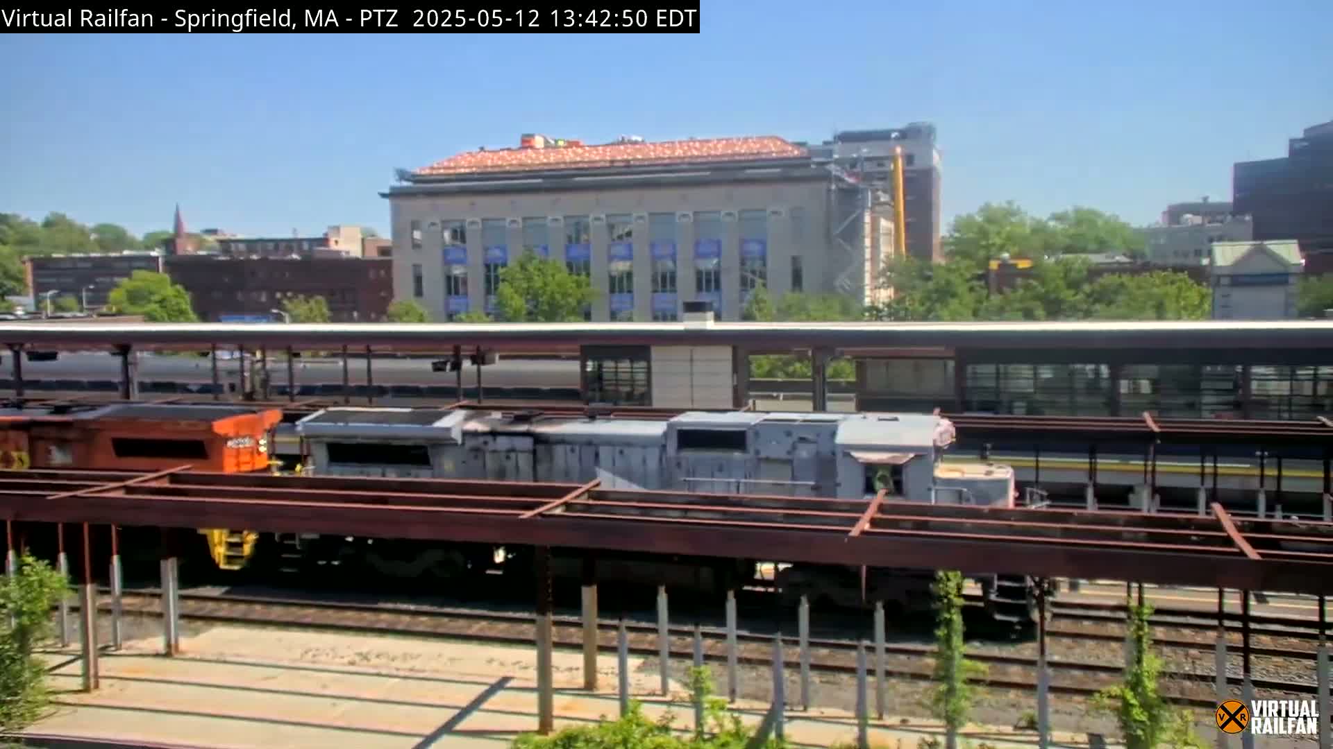 Under a sunny sky, several train cars sit on tracks next to a train platform and a multi-story building undergoing construction.