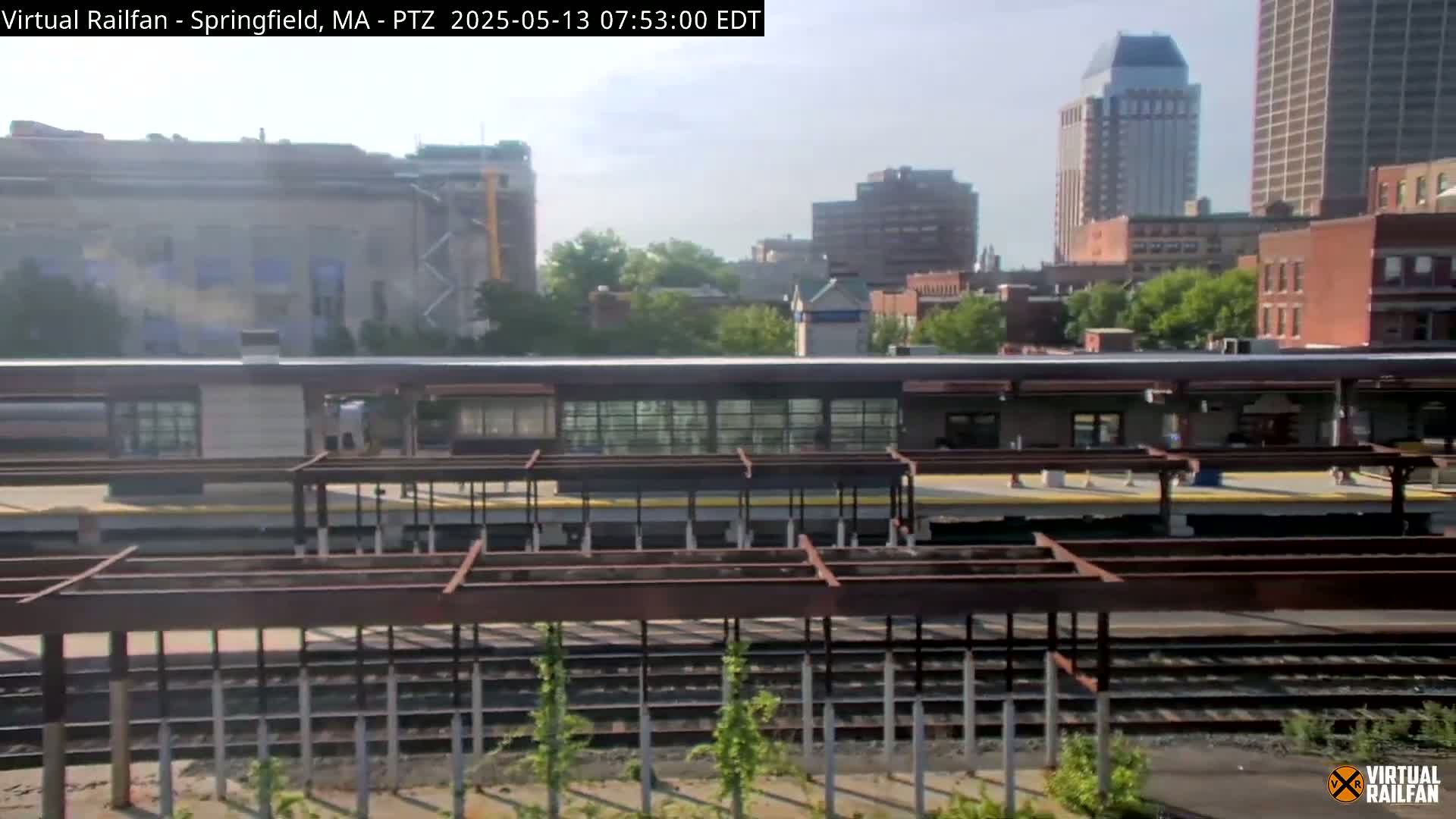 A train station platform with a train partially visible, under a partly sunny sky, is situated amidst a cityscape of several buildings, some under construction.