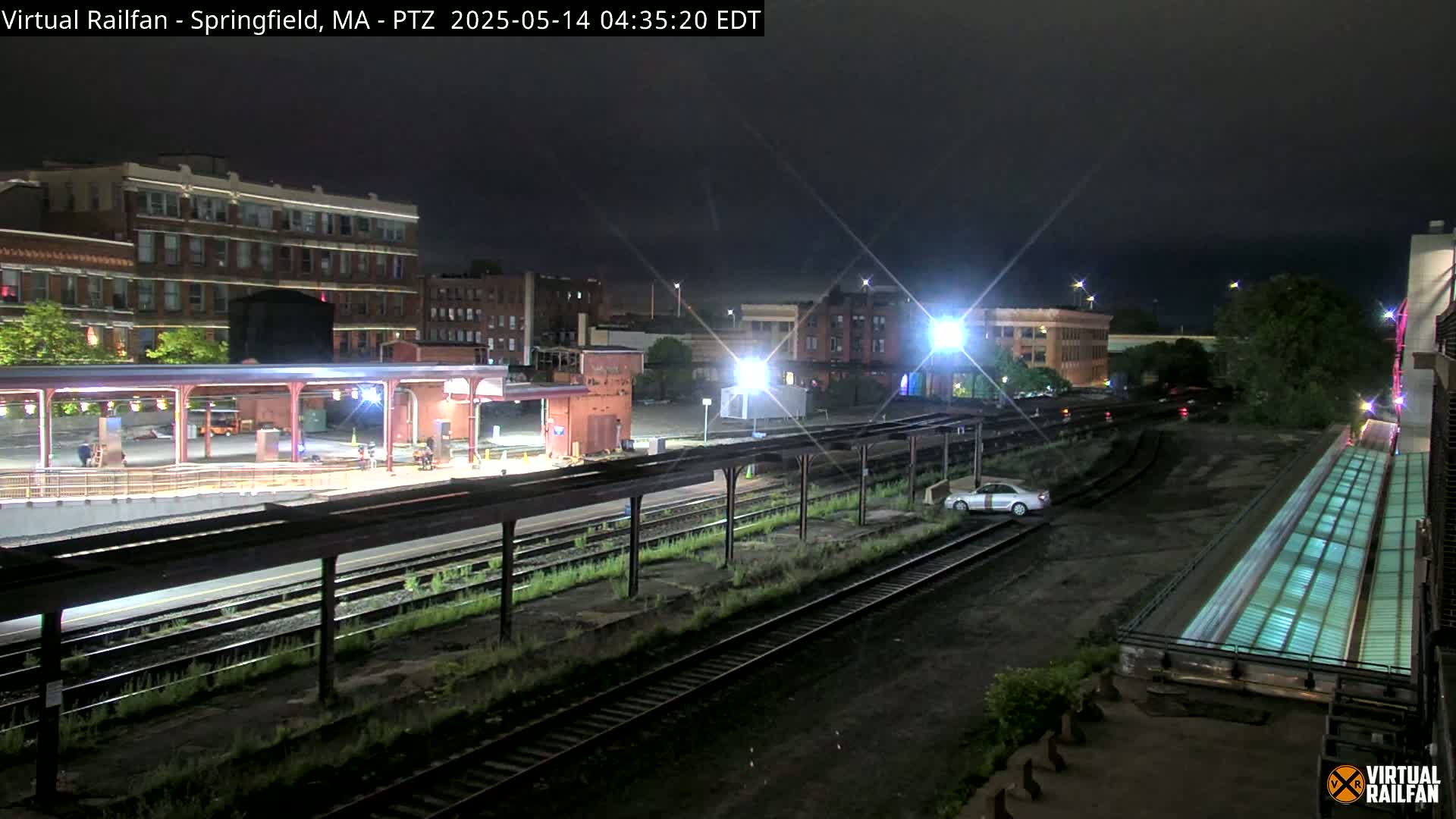 A nighttime view of a train station platform and surrounding buildings, with train tracks and a single car visible in the distance under a dark sky.