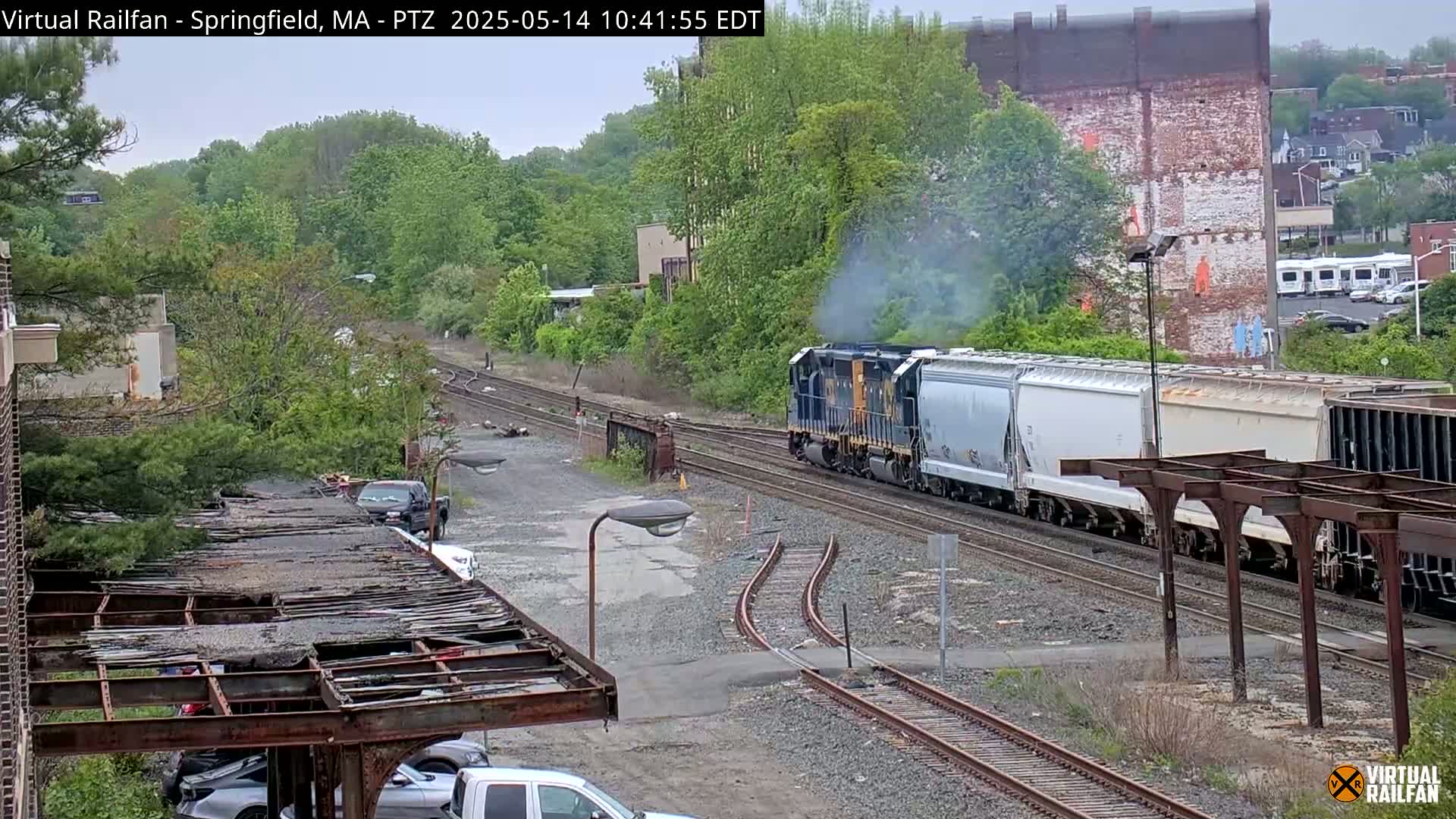 A long freight train, emitting smoke, travels along train tracks surrounded by lush green trees and overcast skies.