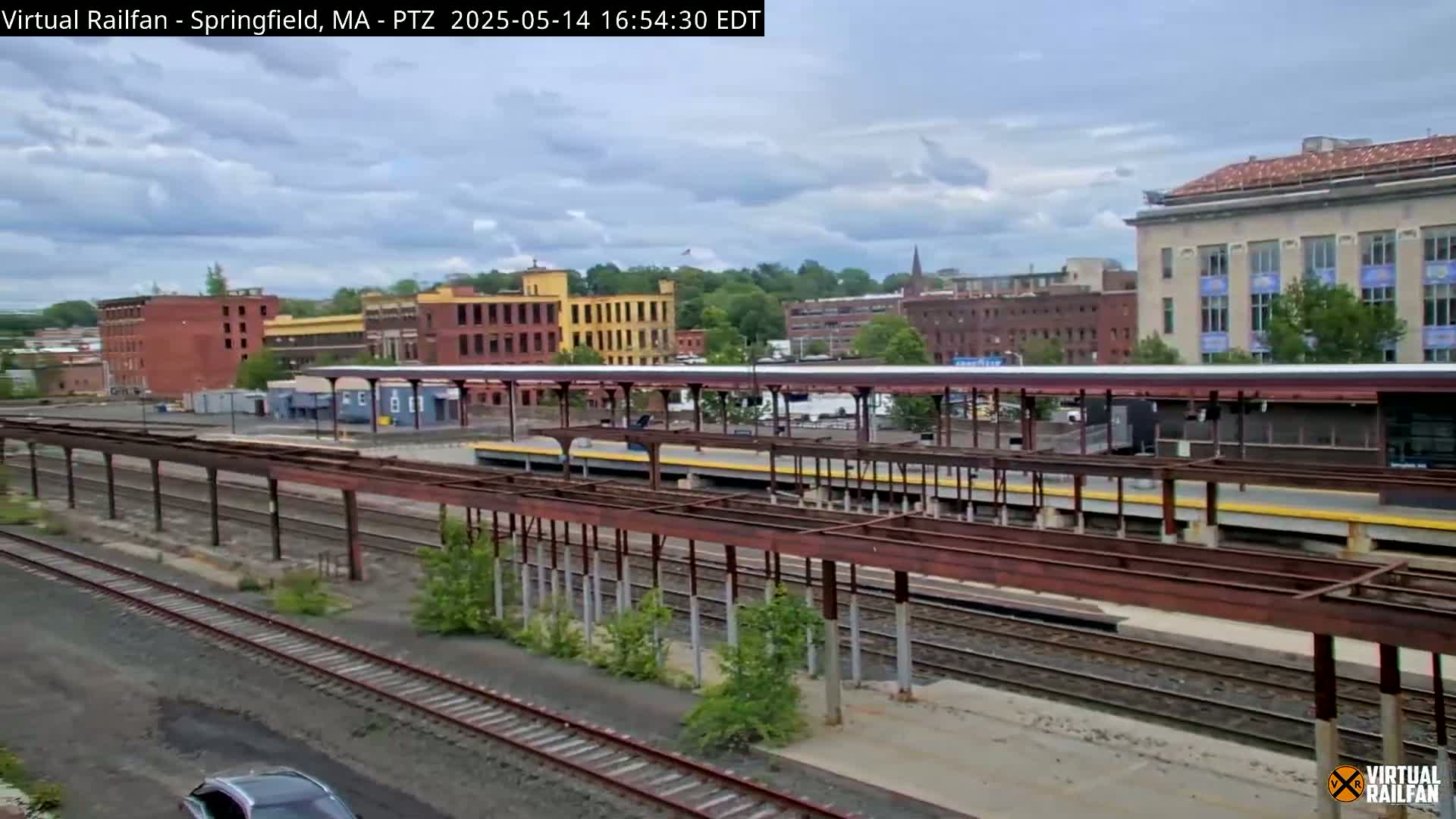 A mostly cloudy day reveals a train station with multiple tracks and platforms, surrounded by industrial buildings and some greenery.