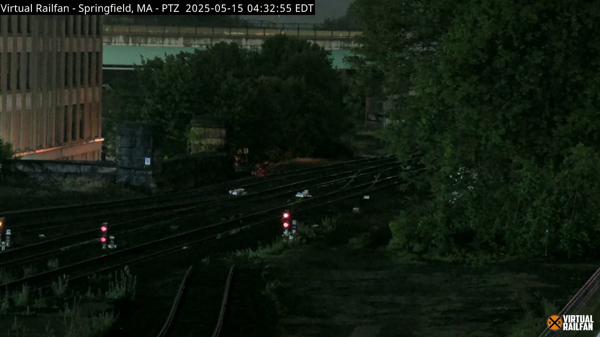 A nighttime view of railroad tracks and signals, partially obscured by overgrown vegetation, near a multi-story building under a dark sky.