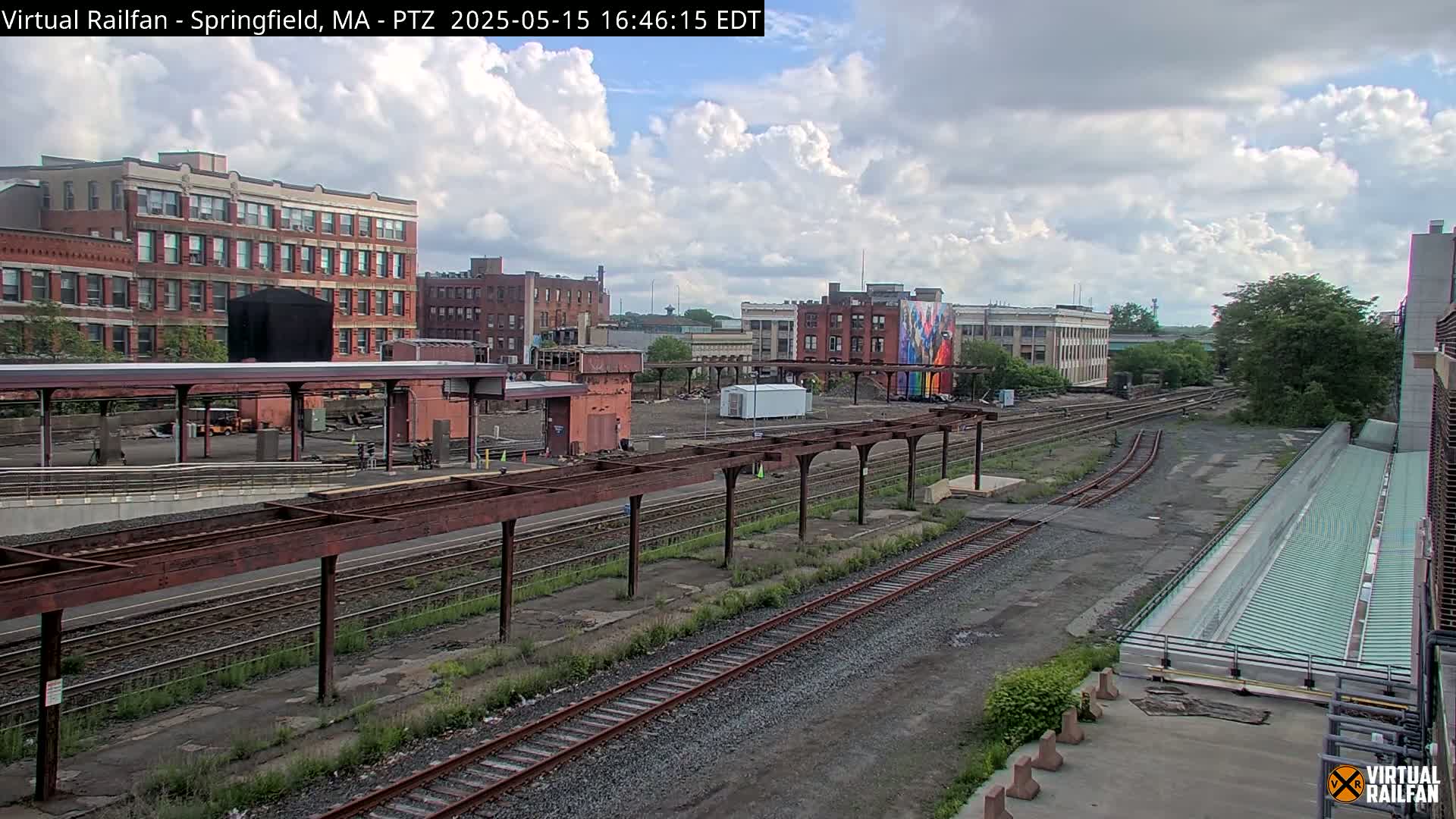 A partly cloudy day reveals a train yard with multiple tracks, railway platforms, and brick buildings in the background.