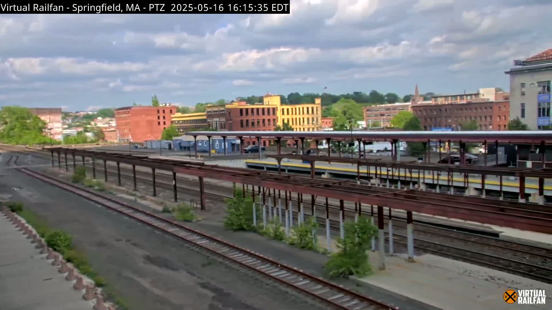 A partly cloudy day reveals a train station with multiple tracks, platforms under covered shelters, and surrounding industrial buildings.