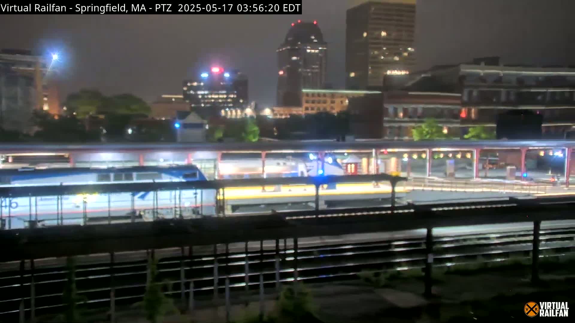 A passenger train sits at a nighttime station, with a city skyline visible in the background under a dark, possibly overcast sky.