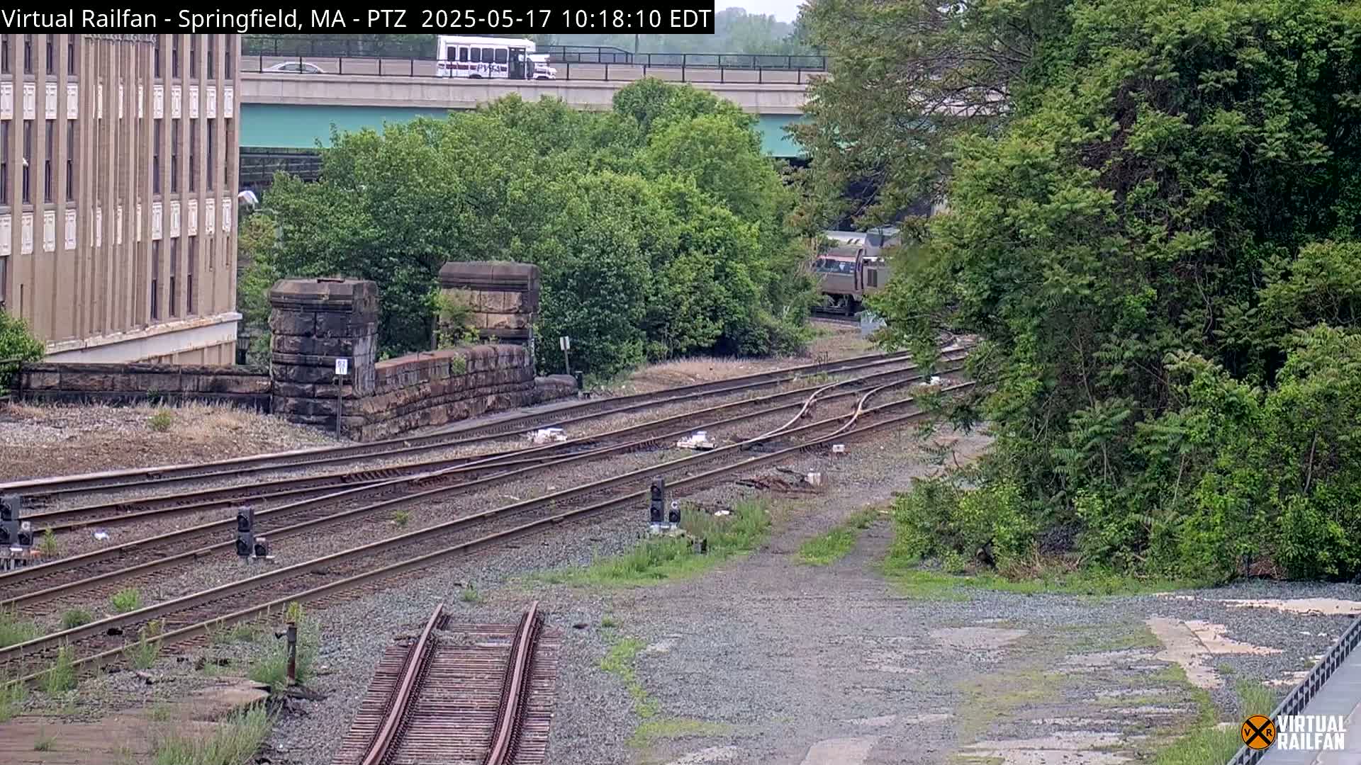 A train passes by a stone wall and multiple railway tracks, under a bridge and next to lush green foliage, on an overcast day.