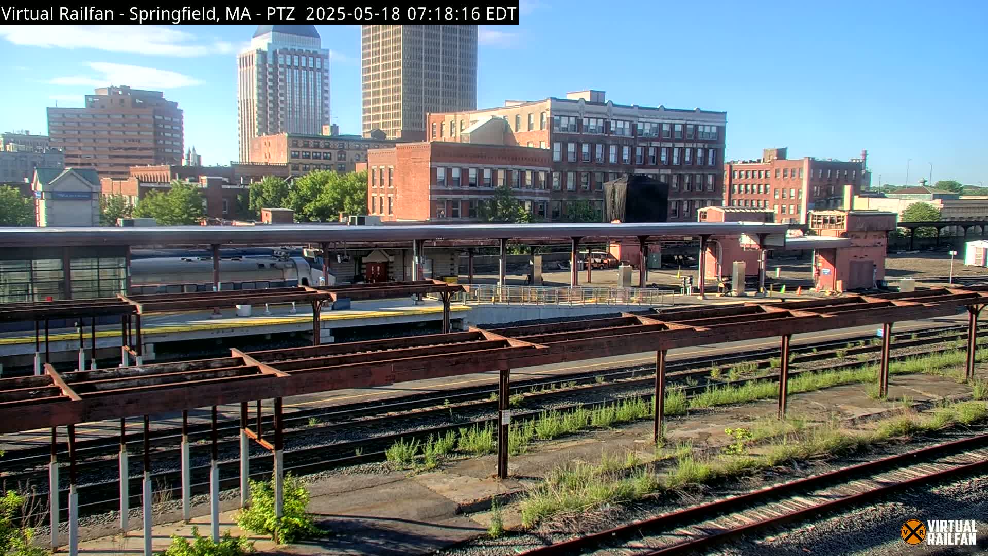 A train sits at a station platform under a covered area, with a city skyline visible in the background on a sunny day.