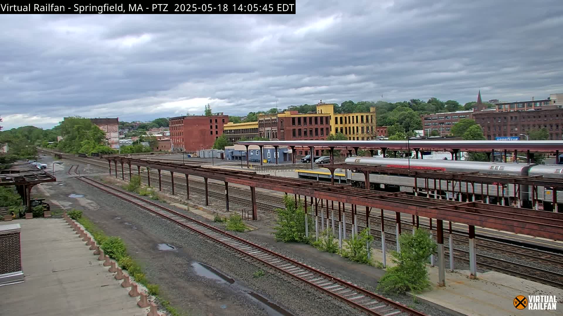 A train sits at a station under an overcast sky, with multiple railway tracks extending into the distance past industrial buildings and greenery.