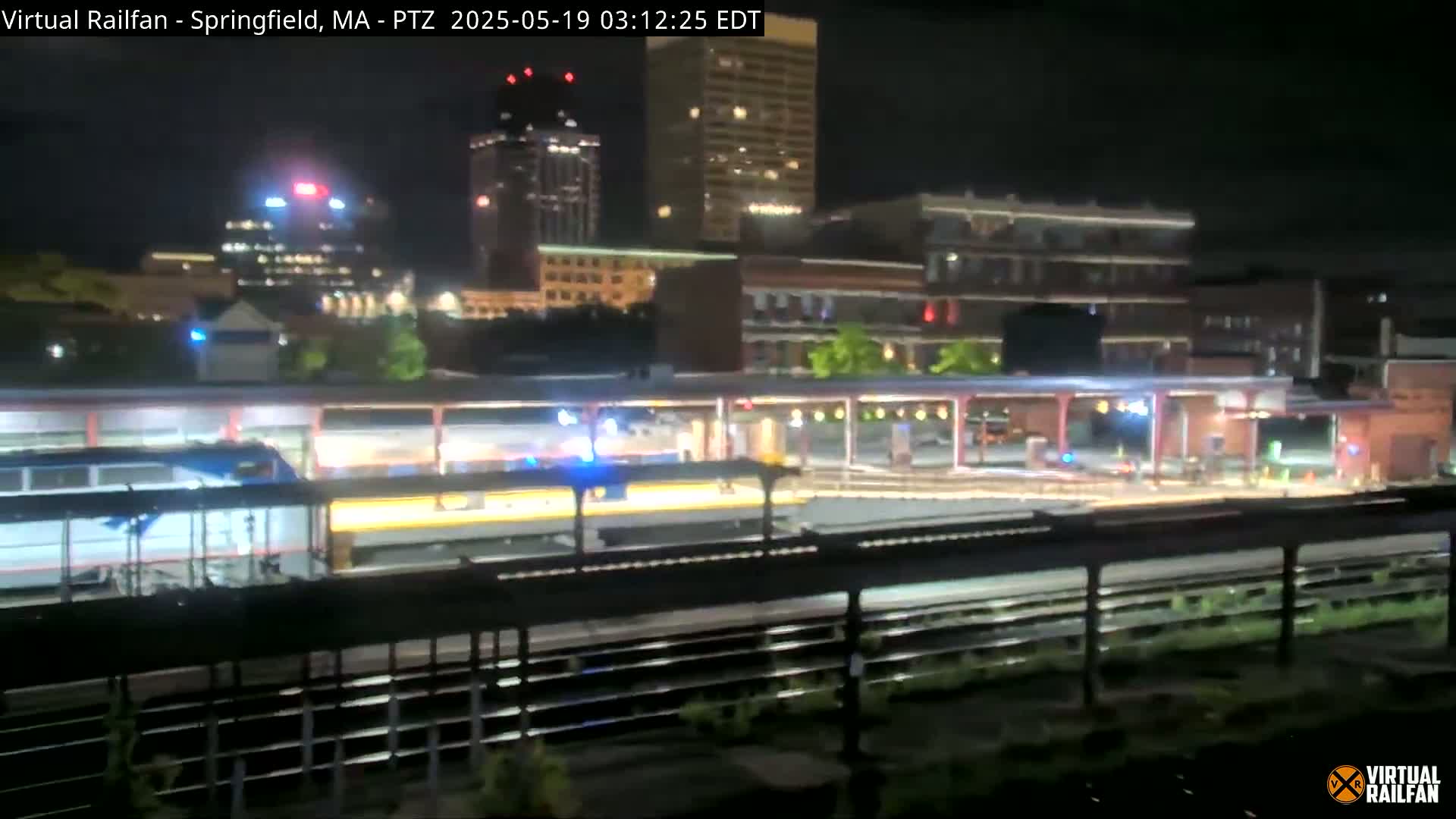 A passenger train sits at a nighttime station in a city, illuminated by artificial lights against a backdrop of buildings.
