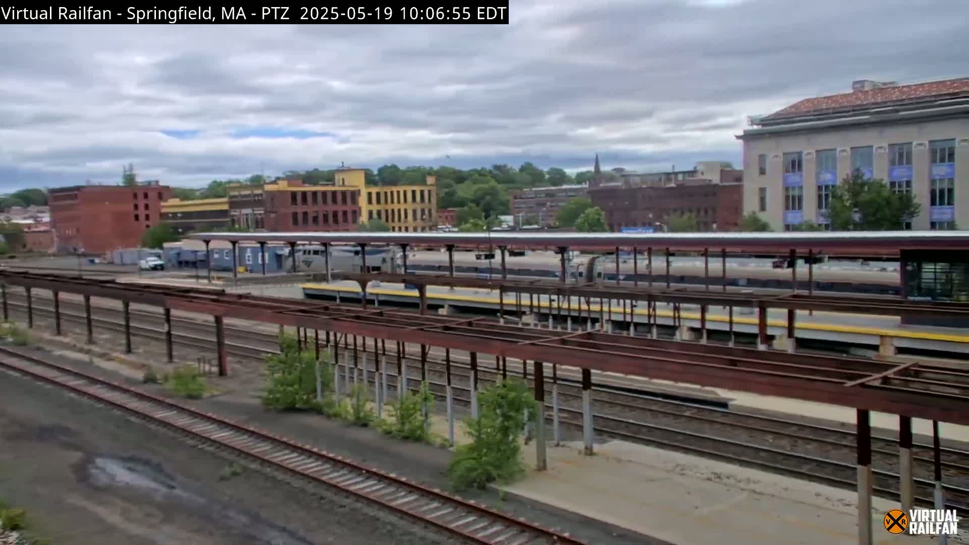 A train station with several train cars under a covered platform, surrounded by industrial buildings under a partly cloudy sky.
