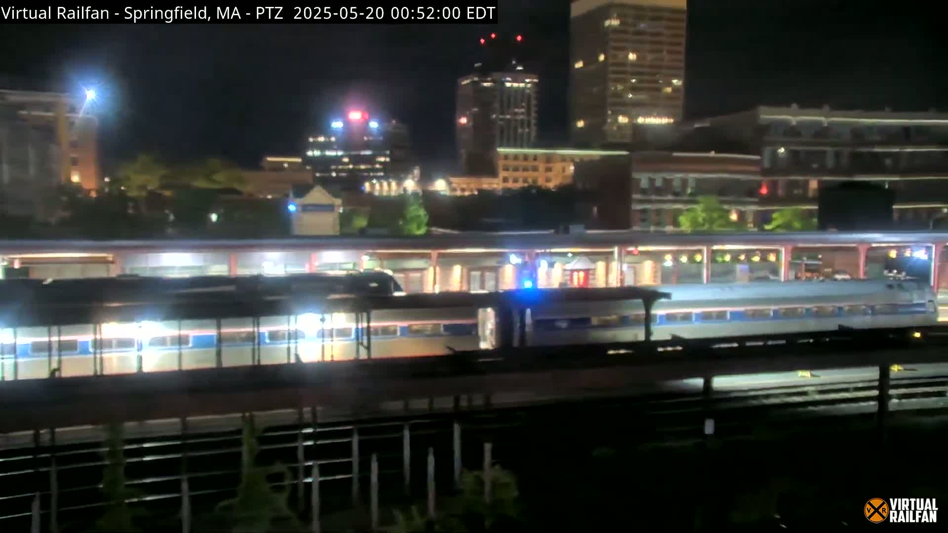 A passenger train sits at a station at night, under a clear sky, with city lights visible in the background.