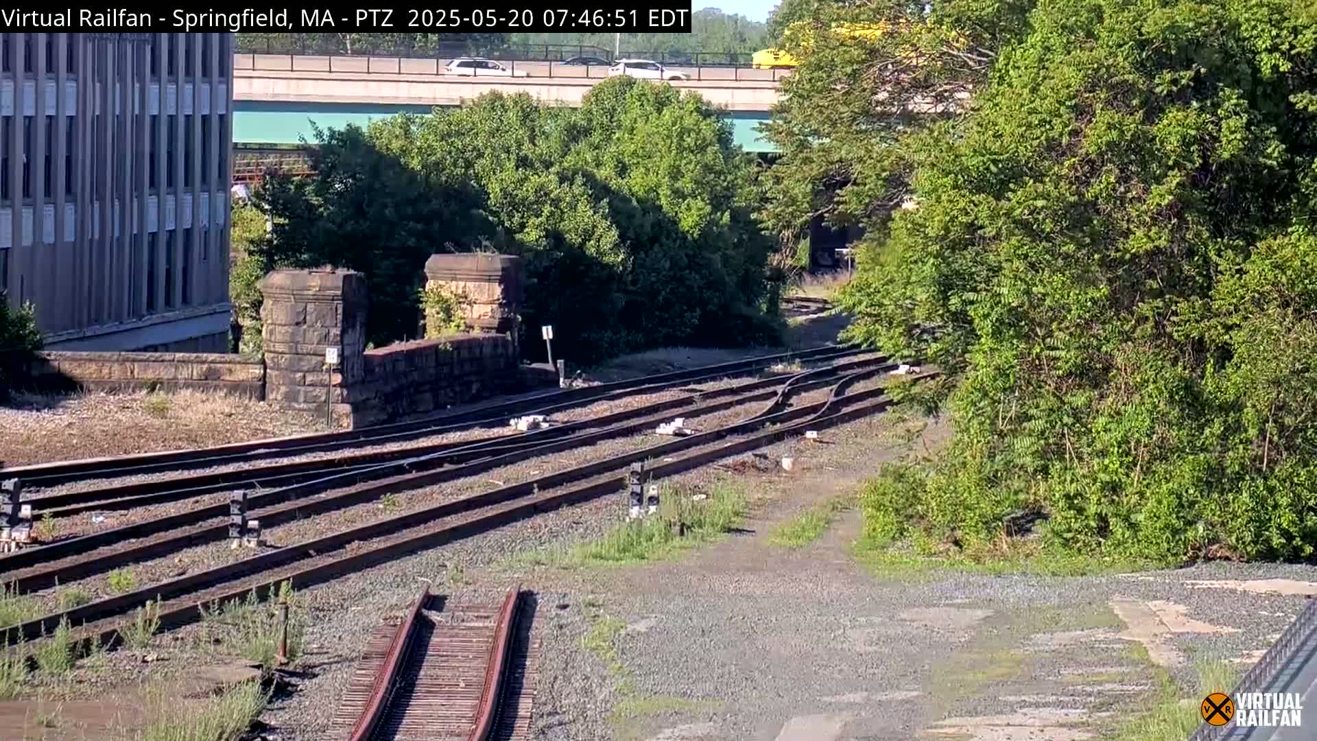 A sunny view of a railway junction with stone retaining walls, lush green vegetation, and a highway overpass in the background.