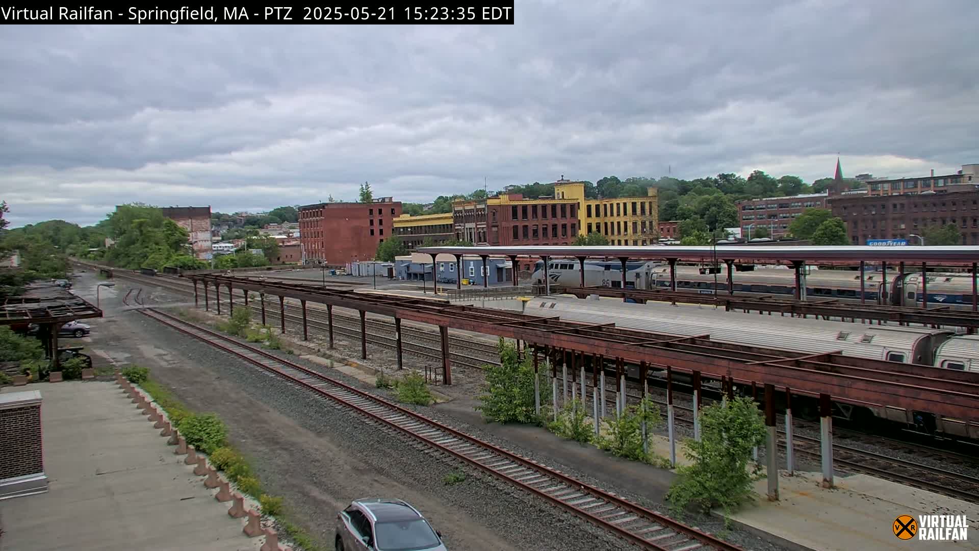 A train station with several passenger cars and surrounding industrial buildings under a cloudy sky.