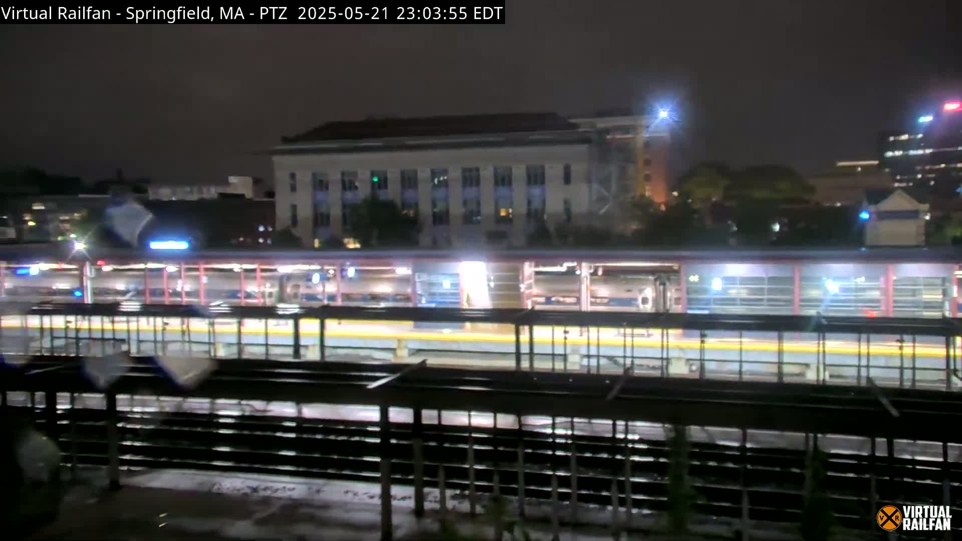 A nighttime view of a train station platform with several trains and a multi-story building in the background.
