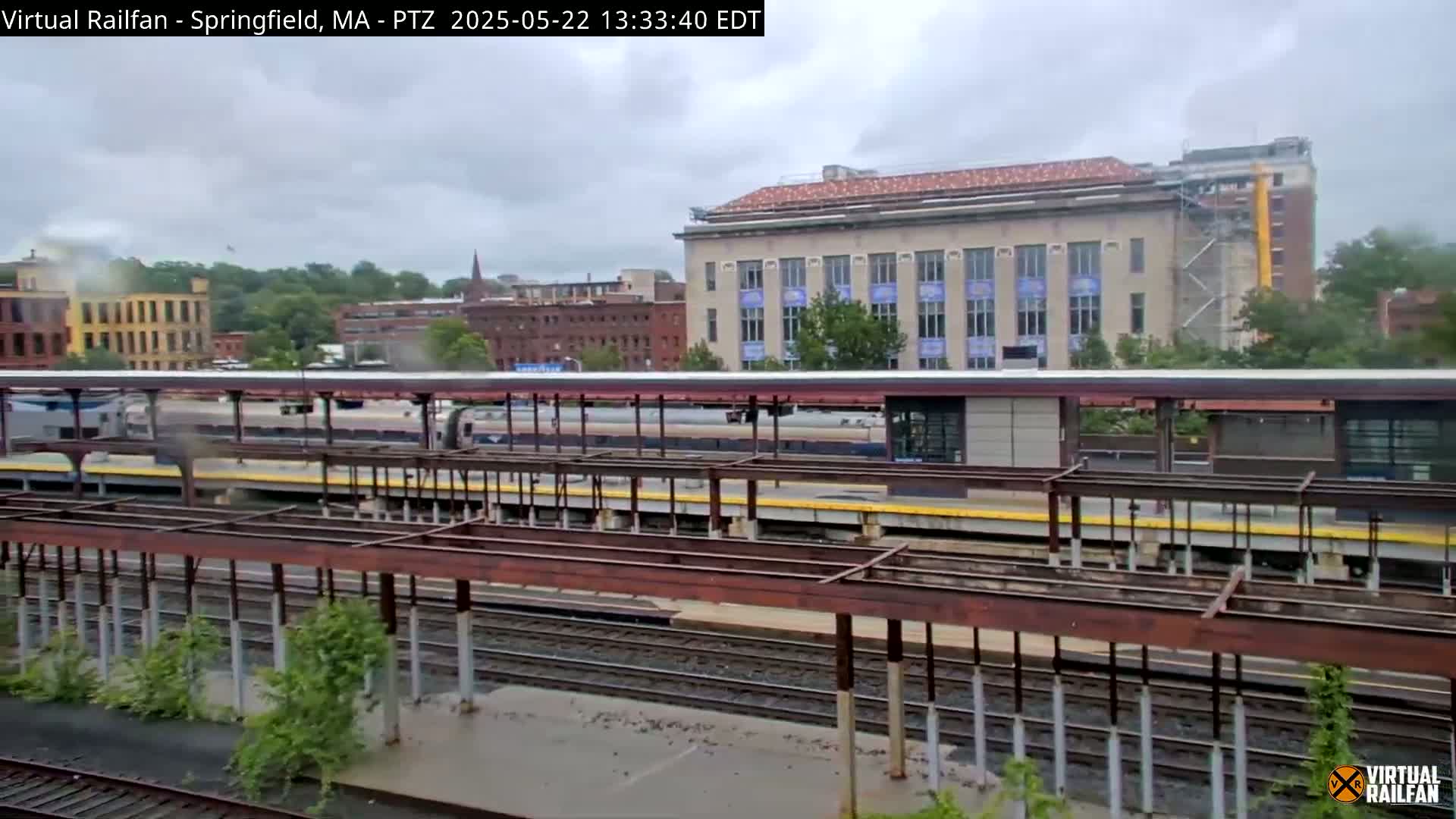 A train sits at a station under an overcast sky, with several buildings visible in the background, one of which is under construction.