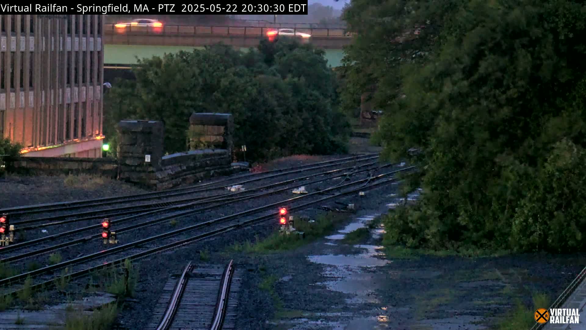 A nighttime view of several railway tracks, some with red signal lights, next to a building and lush green trees, showing some standing water in the ground nearby.