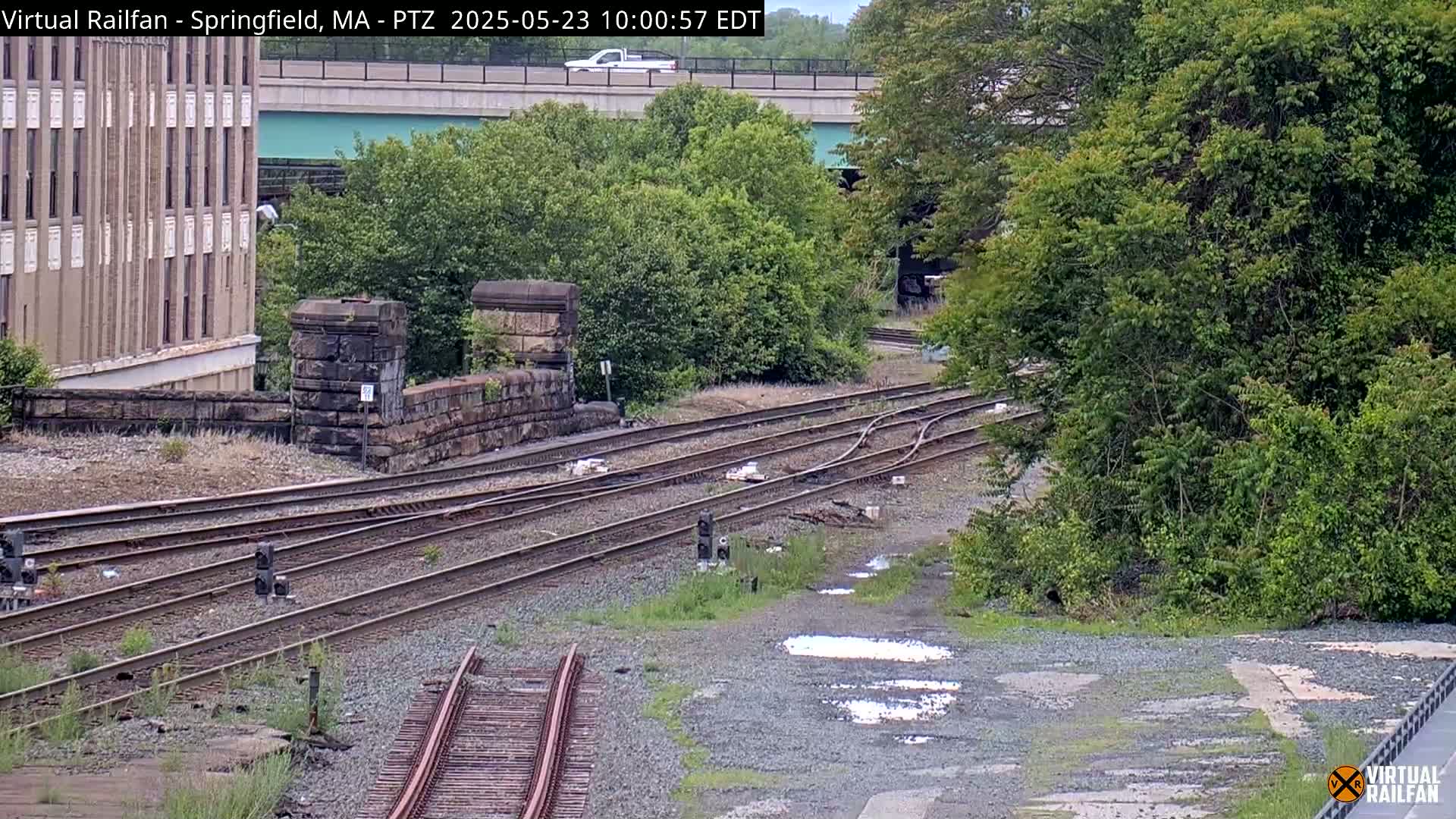 A high-angle view shows several train tracks intersecting near a stone wall and lush green trees under an overcast sky, with puddles of water on the ground near the tracks.