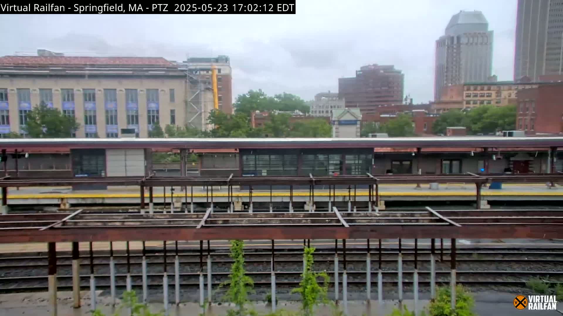 A train station platform with a partially constructed overhead structure is seen under an overcast sky.