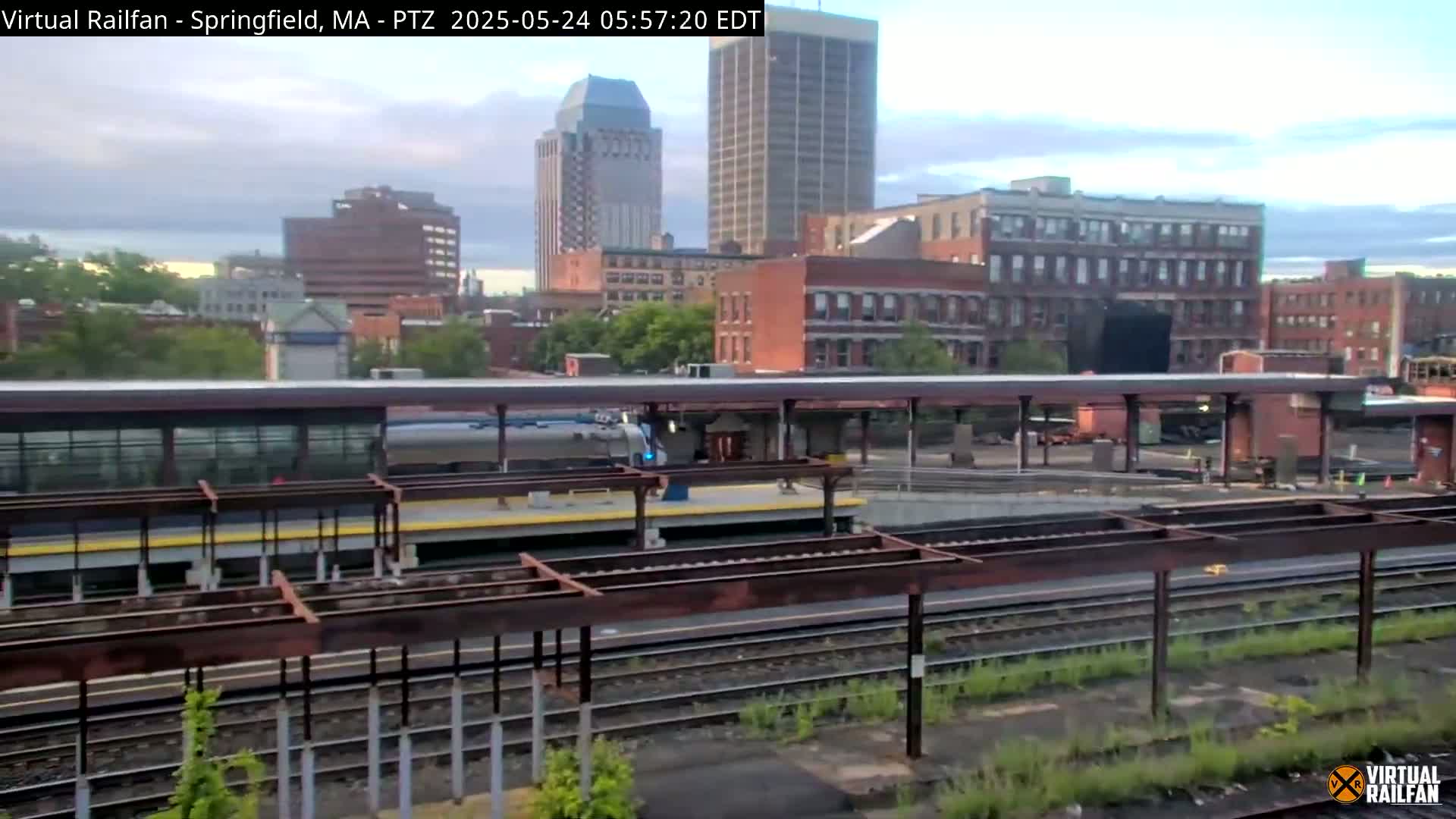 A train sits at a station platform in front of a city skyline under a partly cloudy sky.