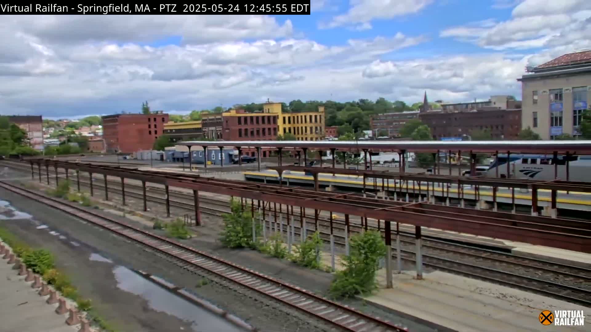 A partly cloudy day shows a train station with multiple train tracks, a passenger train, and industrial buildings in the background.
