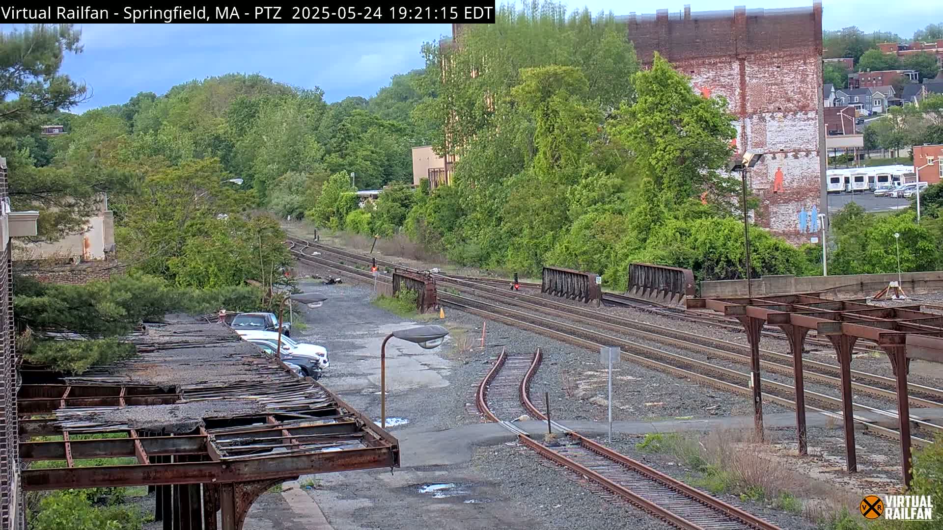 A partly cloudy day reveals several sets of train tracks branching off from each other, surrounded by overgrown vegetation, dilapidated structures, and a large brick building with painted figures on its side.