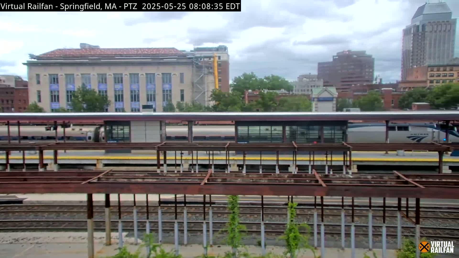 A train sits at a station under an overcast sky, with a cityscape in the background.