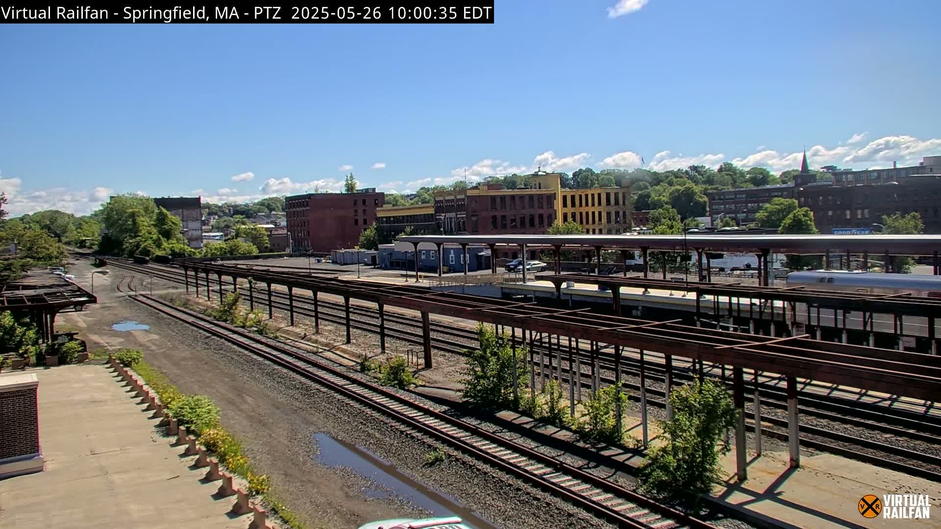 A sunny day reveals a train station with multiple train tracks, a platform covered by a rusty metal structure, and several industrial buildings in the background.
