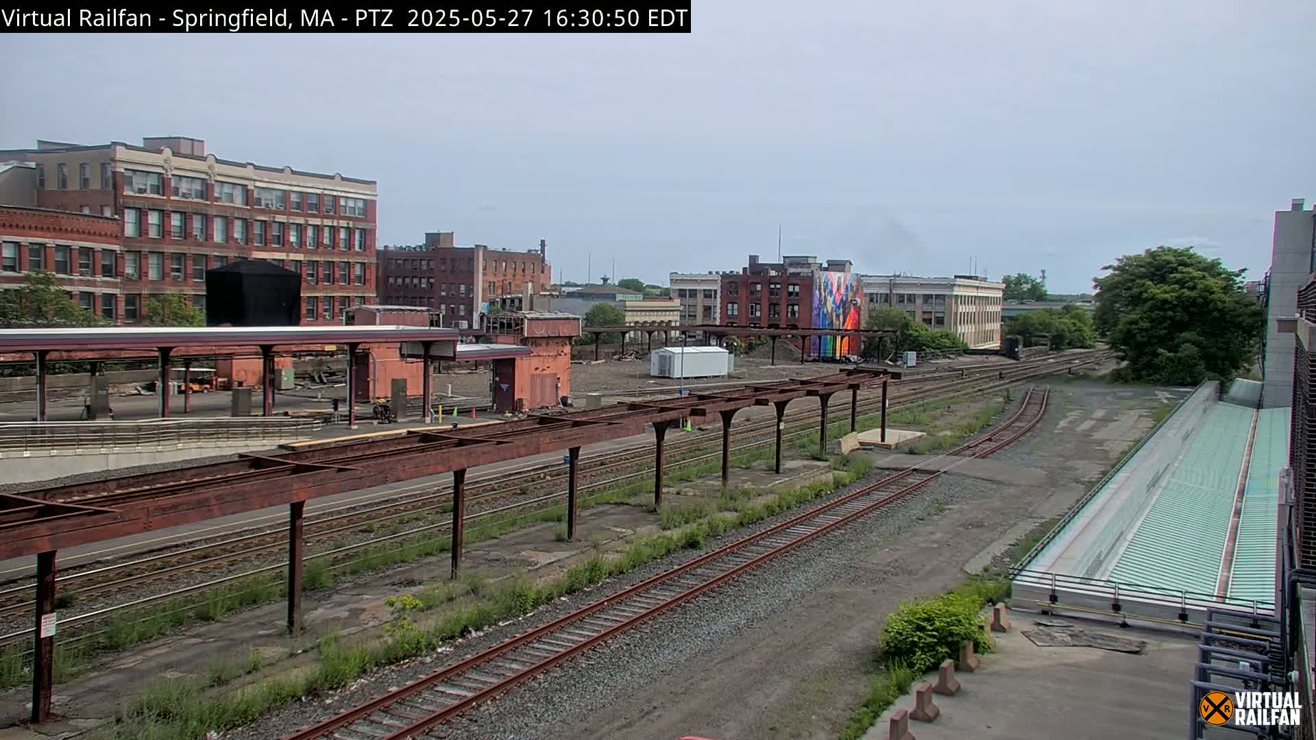 A view of a train station under a partly cloudy sky, showing railway tracks, platforms, and brick buildings in the background.