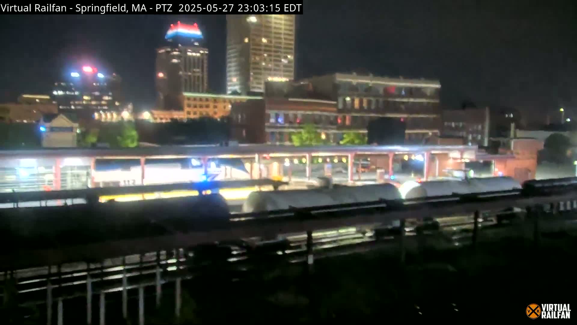 A nighttime view of a train at a station in front of a city skyline.