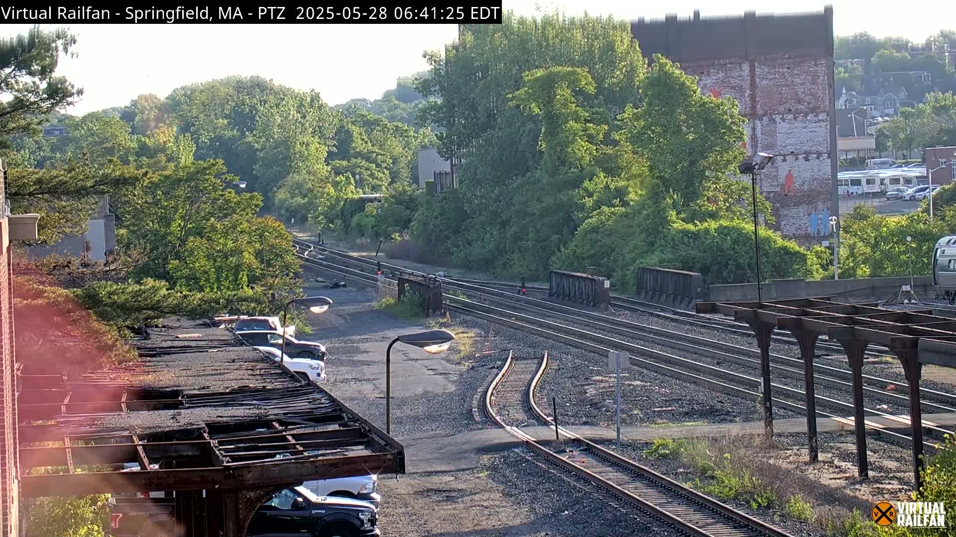 A sunny view of a railroad yard with several sets of tracks, surrounded by lush green trees and buildings, including a brick building with a mural.