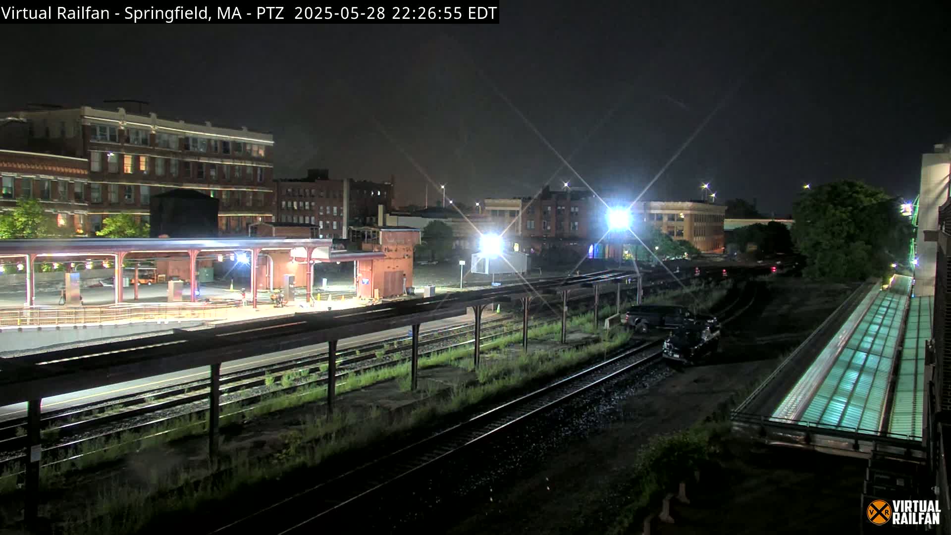 A nighttime view of a train station platform and railroad tracks, with several buildings visible in the background under clear skies.
