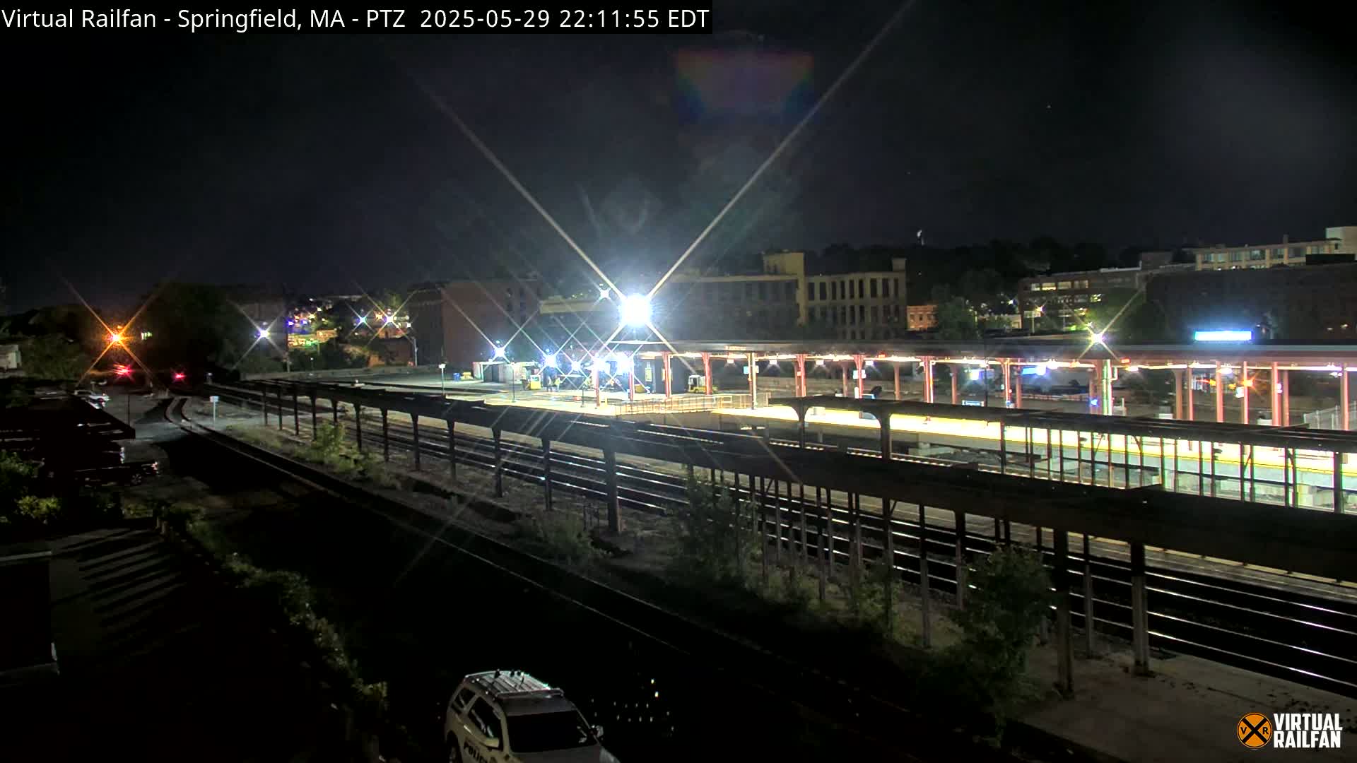 A nighttime view of a train station platform with multiple train tracks, illuminated by bright lights, under a clear sky.