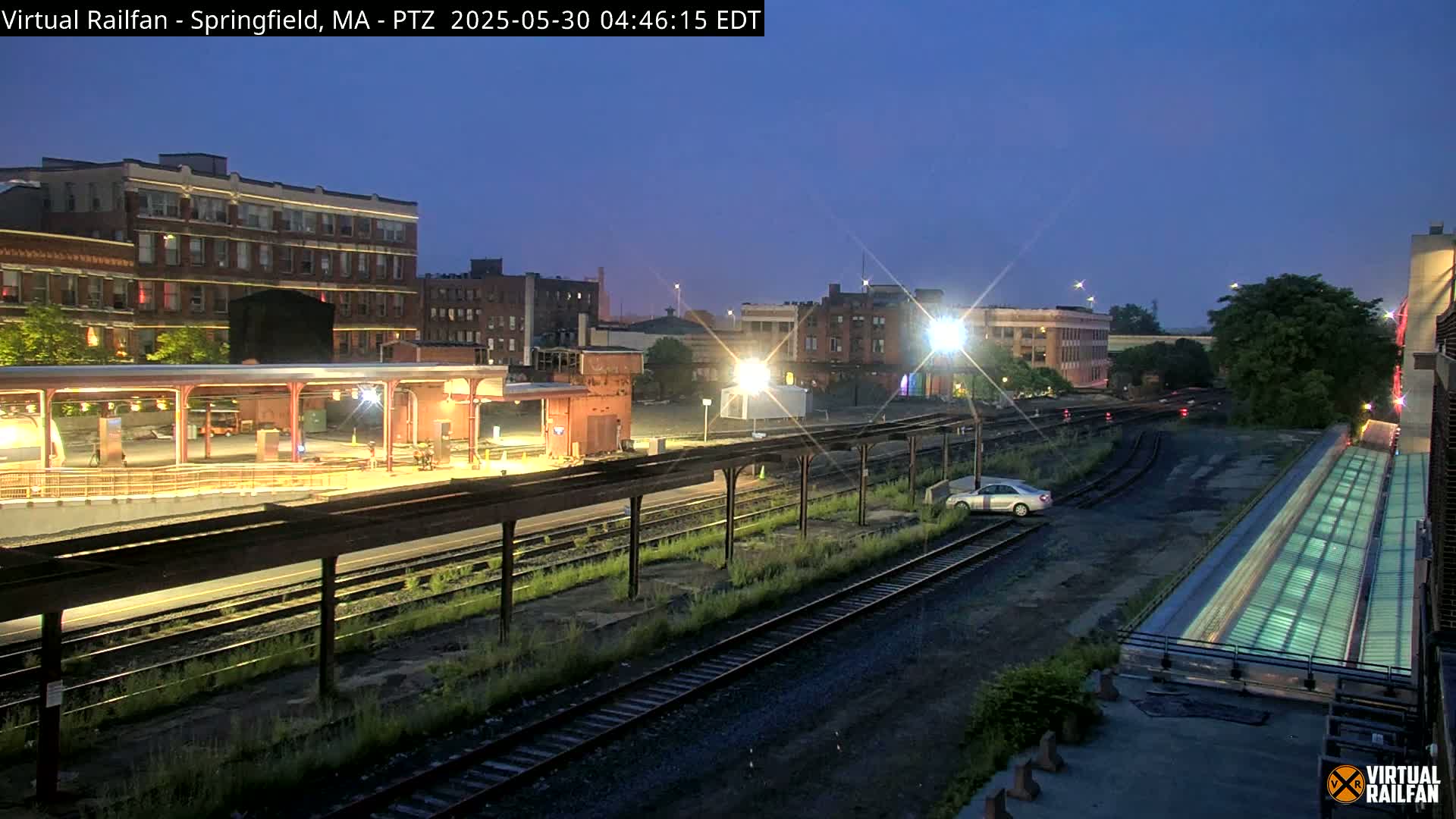 A nighttime view of a train station with a few buildings in the background, under a twilight sky.