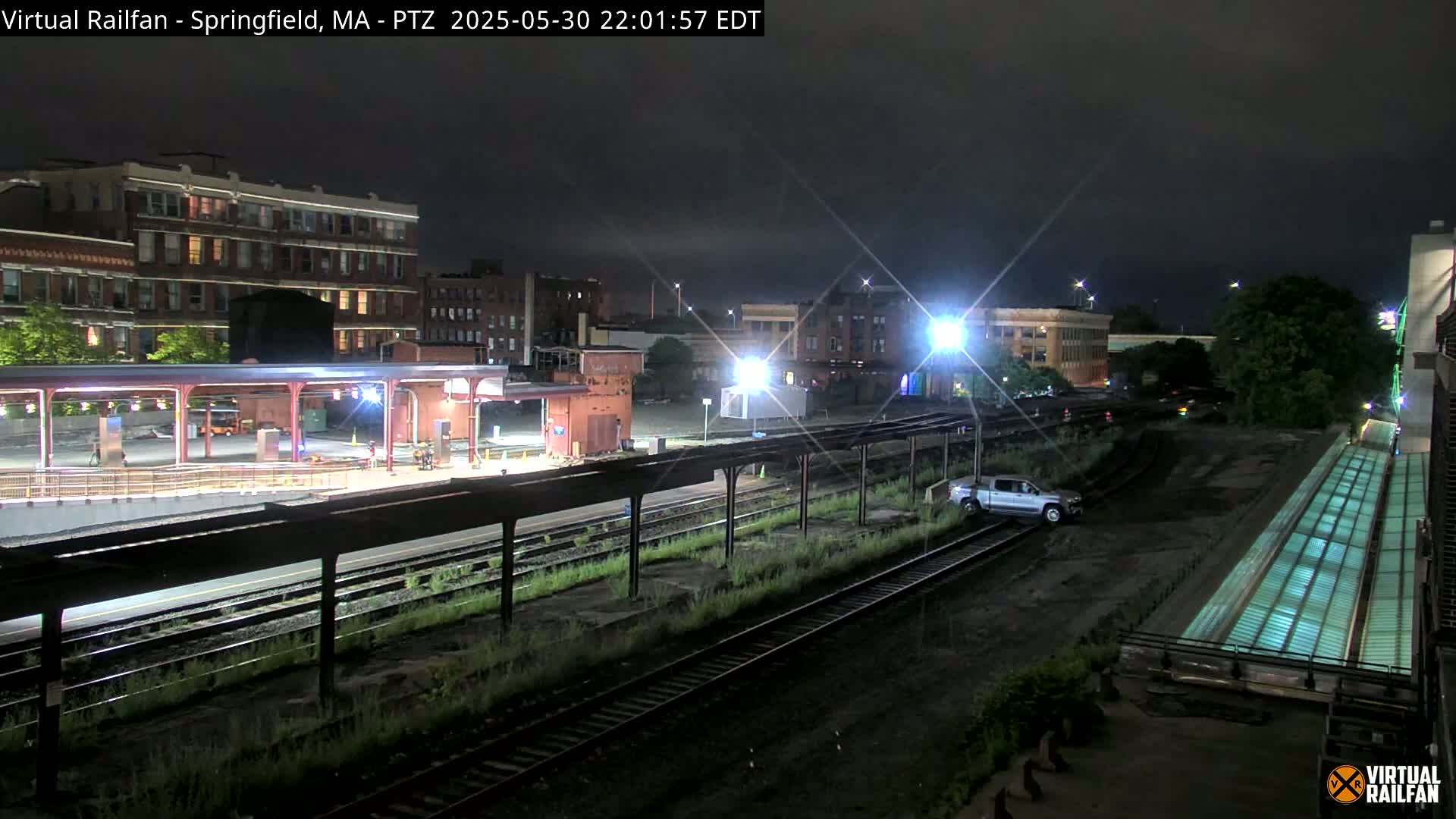 A nighttime view of a train station platform and tracks, with buildings in the background under a dark, clear sky.