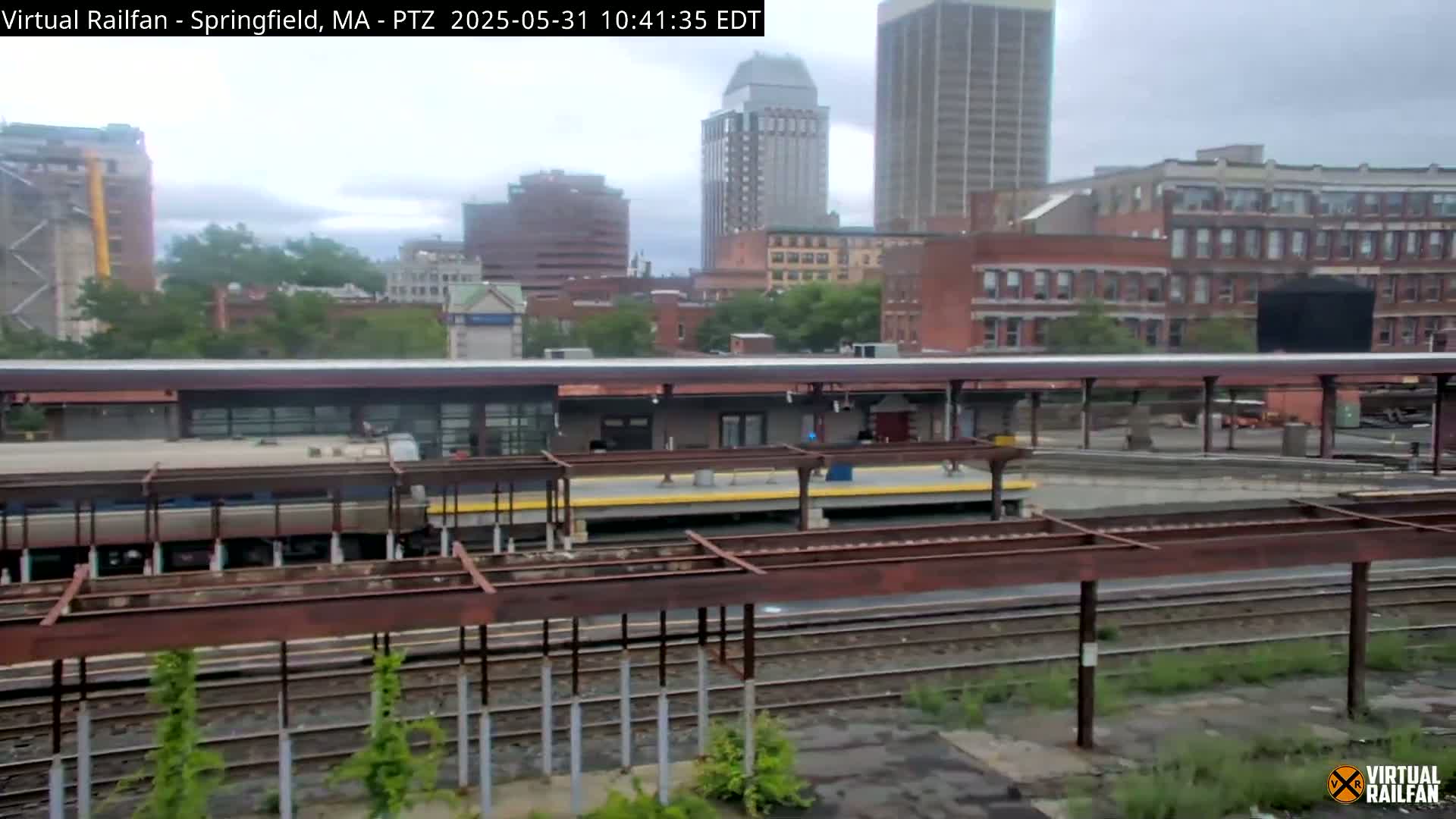A train station platform with a train partially visible, under an overcast sky, situated among city buildings.