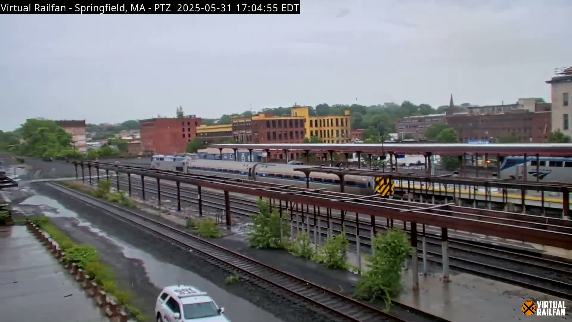 A long passenger train sits at a station on a rainy day, with several industrial buildings visible in the background.
