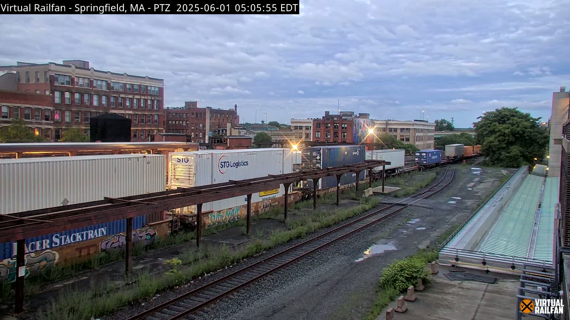 Under a cloudy twilight sky, a freight train with several shipping containers sits on railroad tracks near brick buildings.