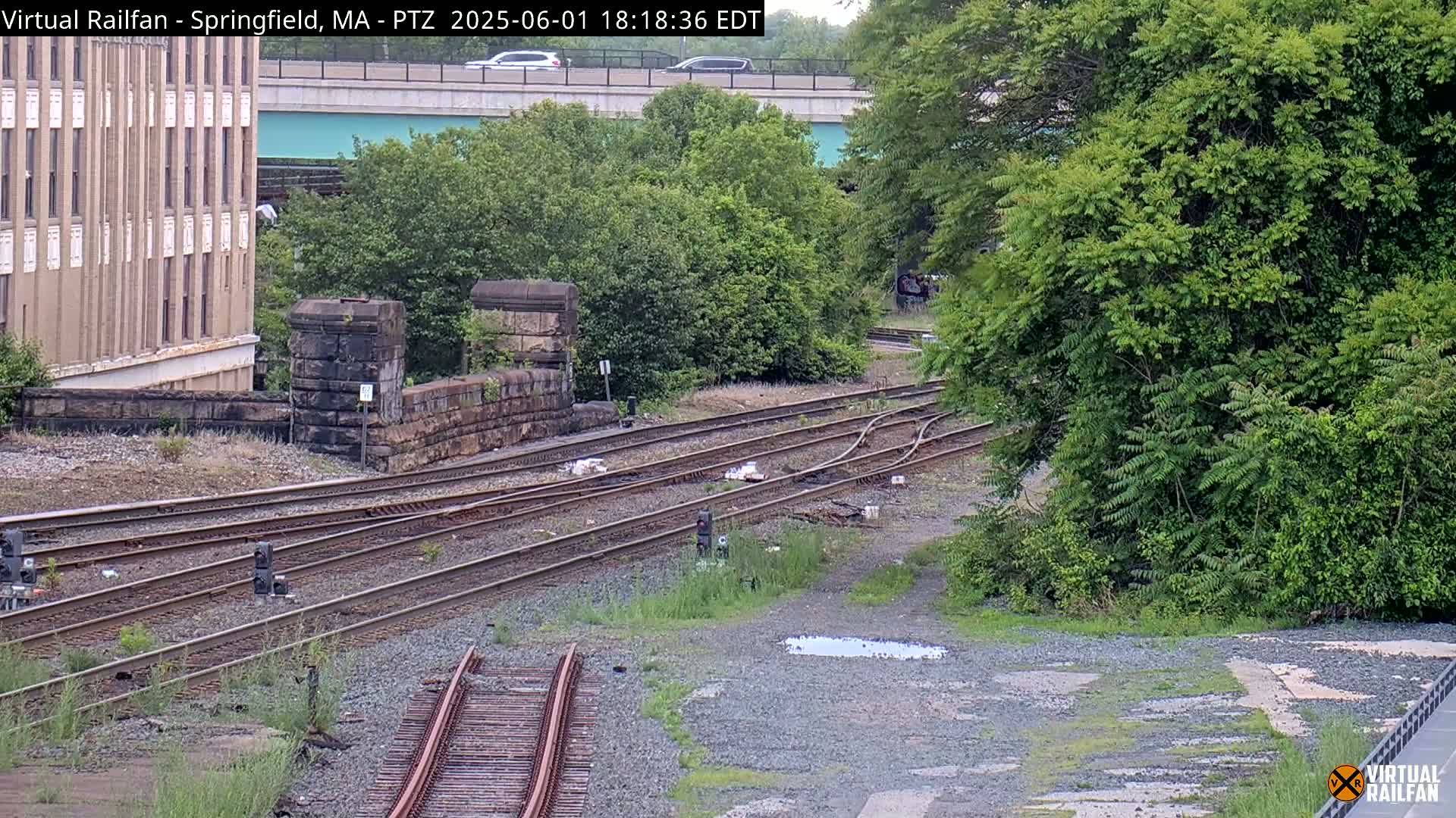 Several sets of train tracks intersect near a stone wall and a multi-story building under an overcast sky.