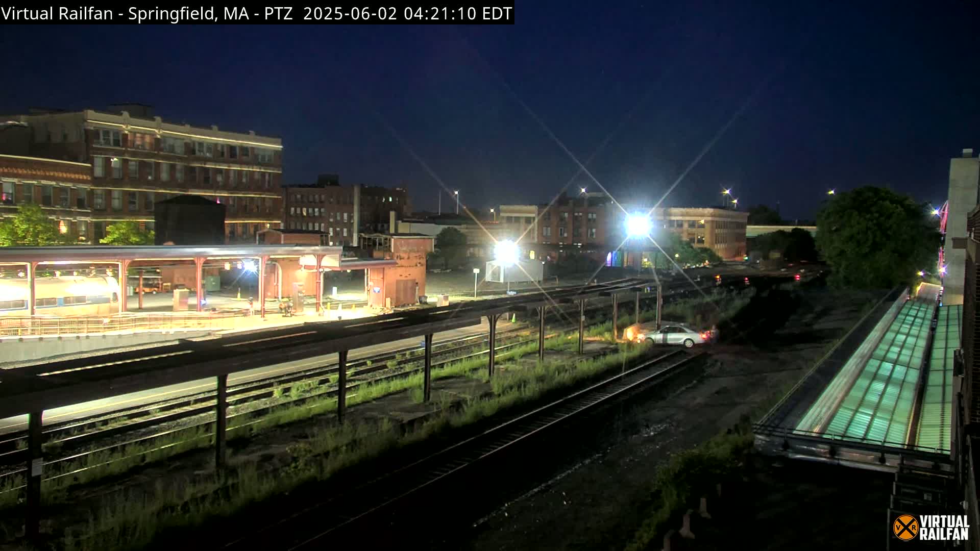 A nighttime view of a train station with a train car visible, a car parked near the tracks, and buildings in the background under a clear, dark sky.