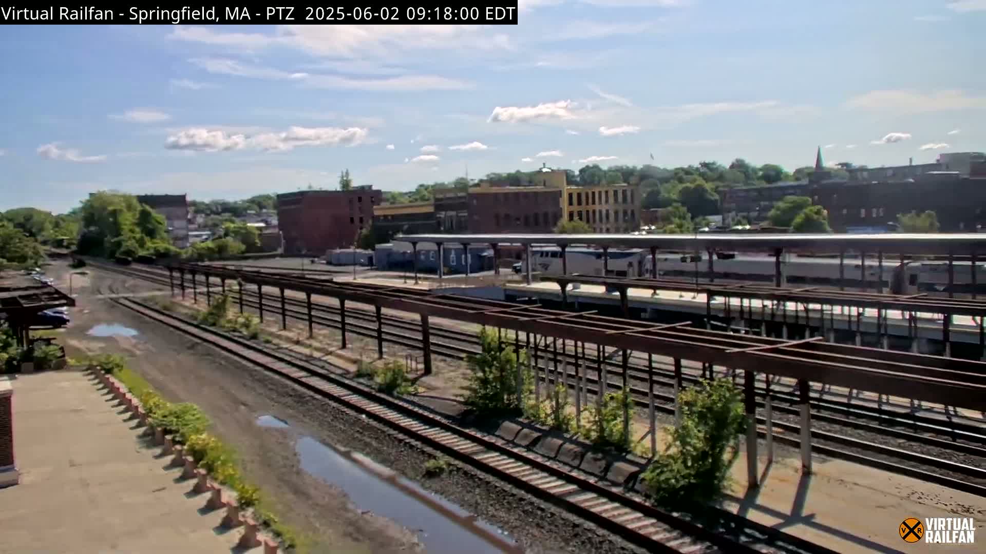 A mostly sunny day reveals a train station with several train cars at the platform and multiple railway tracks extending into the distance, alongside industrial buildings and some vegetation.